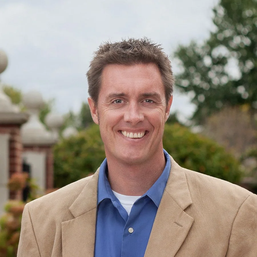 A man with short, dark, spiked hair, smiling, wearing a tan blazer over a blue shirt, outdoors with trees and a building in the background.