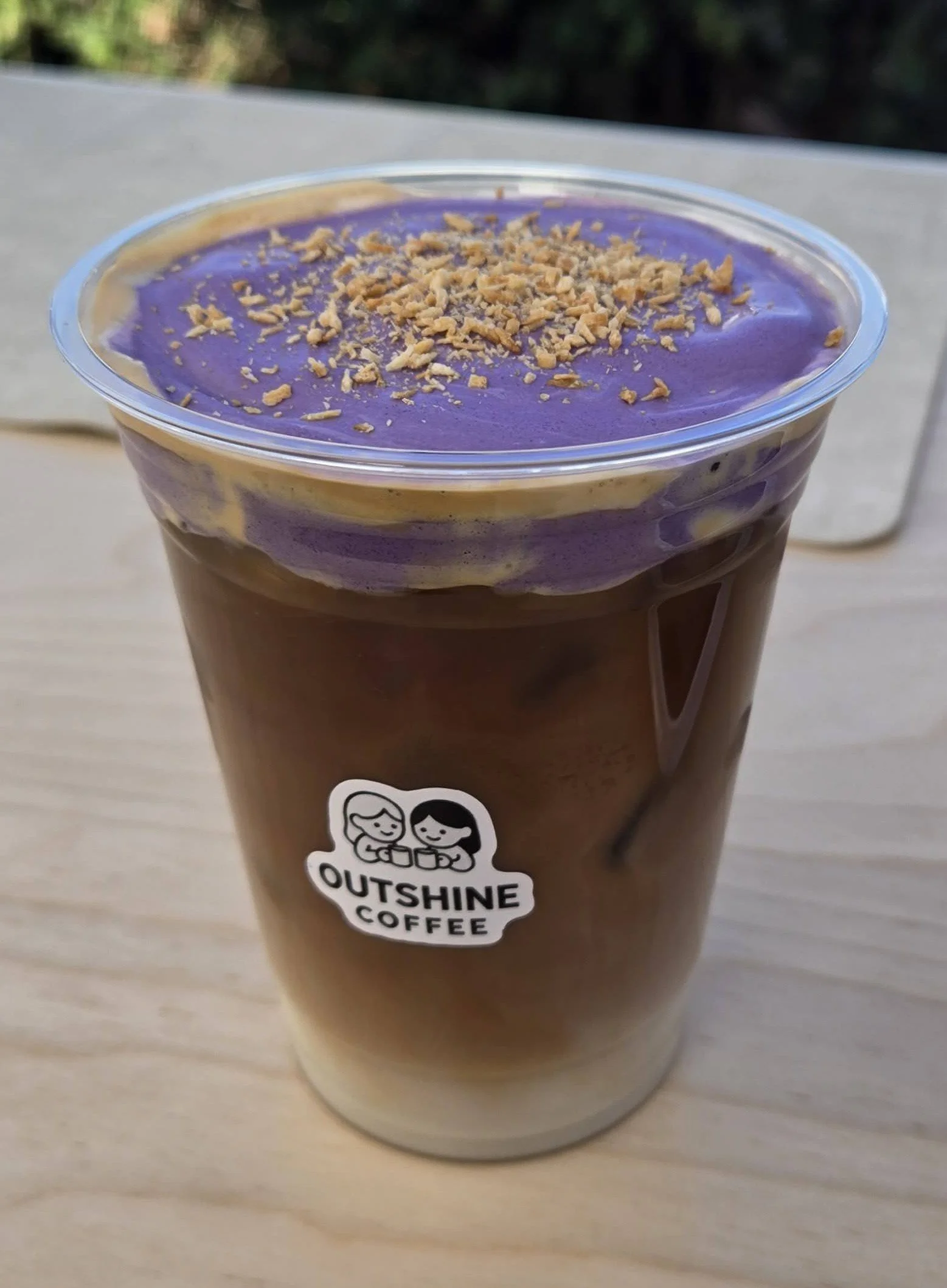 A plastic cup of iced coffee topped with purple and yellow swirled cream and sprinkled with crushed nuts, with a label that says 'Outshine Coffee'.