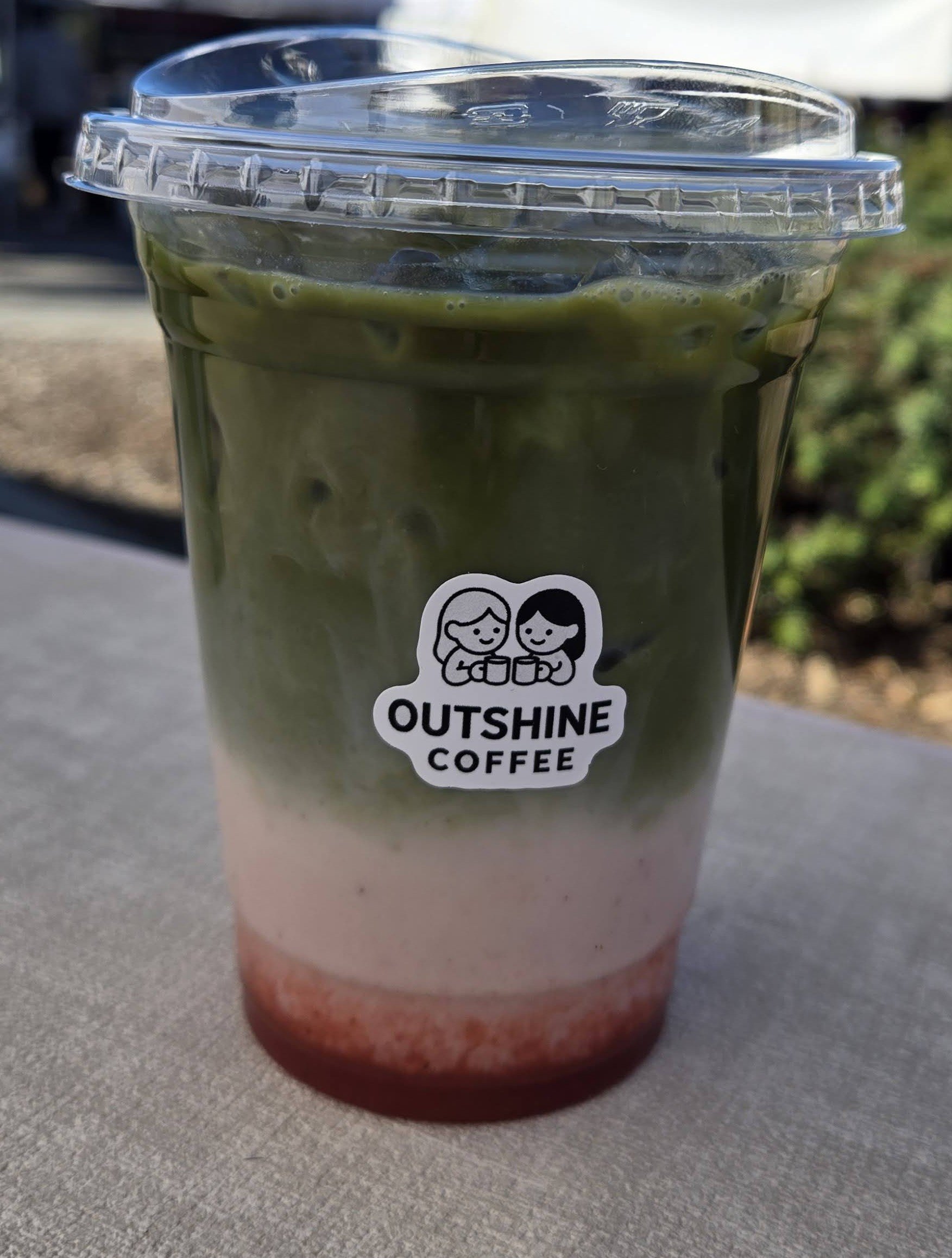 A triple-layered iced beverage in a clear plastic cup with a lid, featuring a white sticker with the OUTSHINE COFFEE logo depicting two cartoon characters holding cups, set on an outdoor table.
