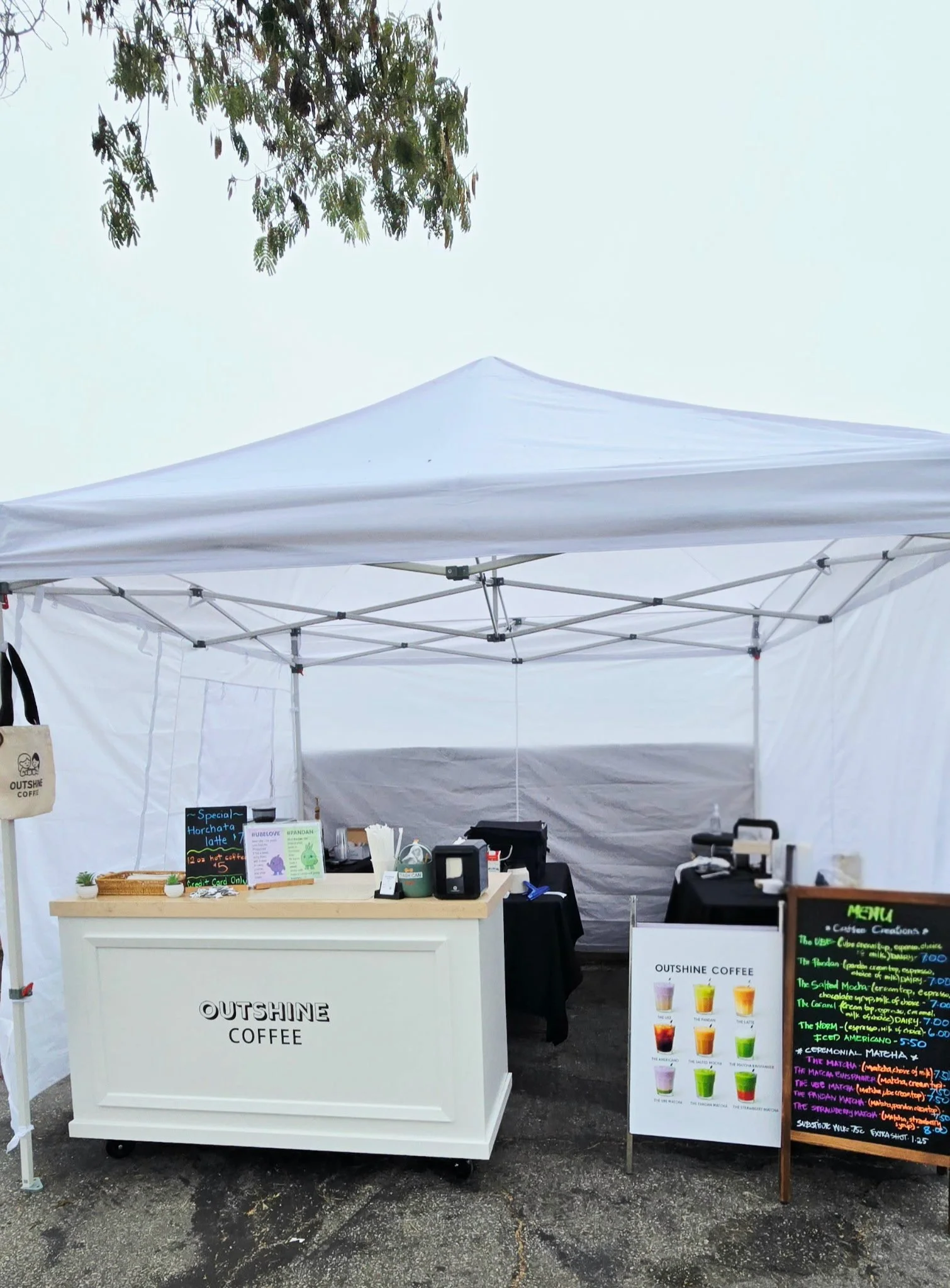 A small outdoor coffee stand named OUTSHINE COFFEE with a white service counter, a menu board, and various items for sale, set up under a white canopy tent on an outdoor surface with a tree branch visible in the upper left corner.