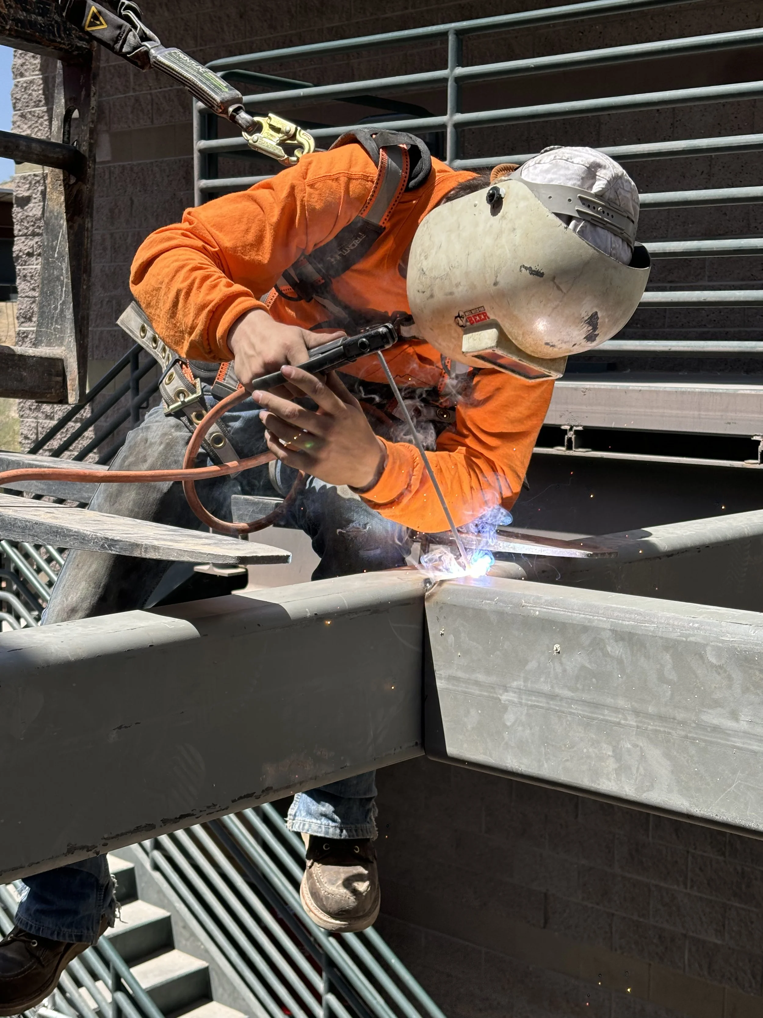 Worker in orange shirt and helmet welding on metal structure outdoors.