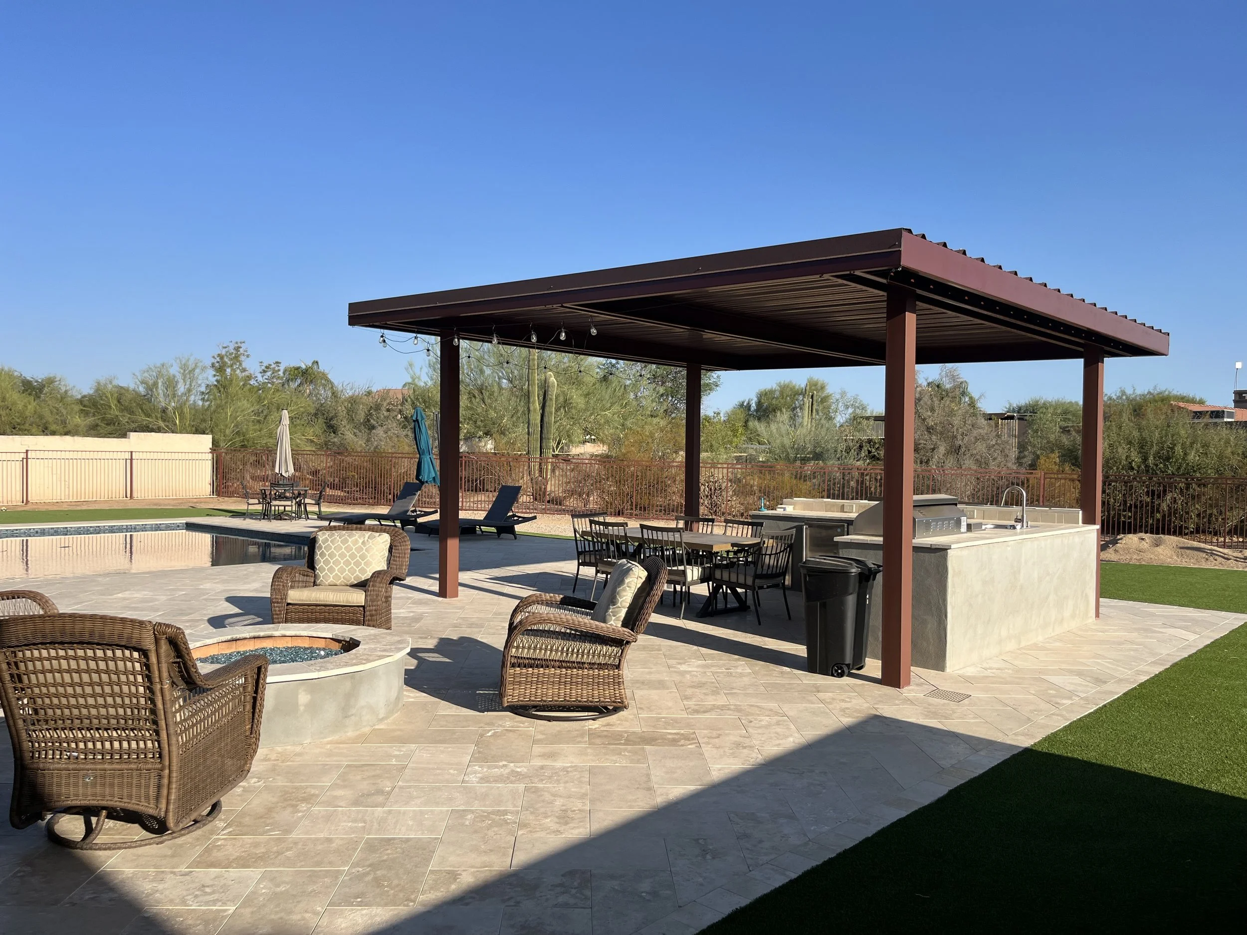 Outdoor pool area with a covered kitchen and seating area, lounge chairs, a fire pit, and a fenced yard with desert landscaping under a clear blue sky.