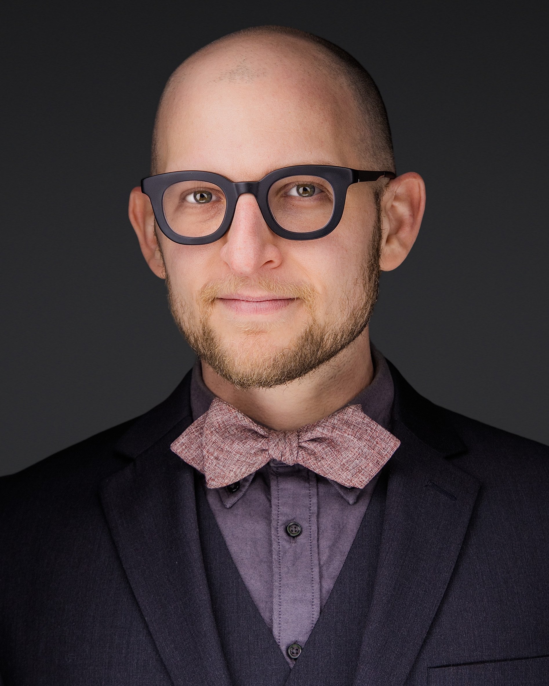 Portrait of a man with glasses, a beard, and a mustache, wearing a suit jacket, shirt, and a bow tie, against a dark background.