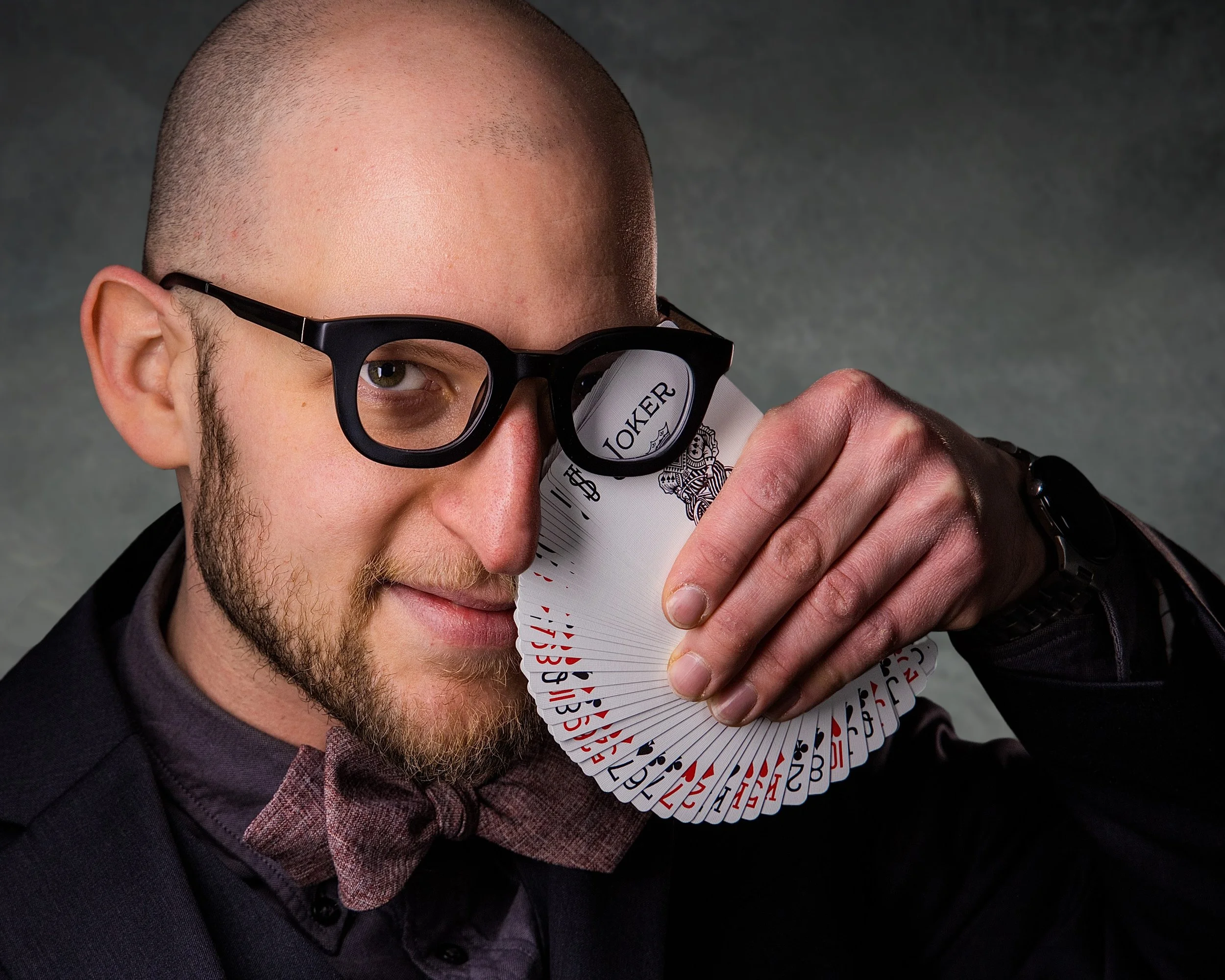 A man with glasses and a bow tie holding a deck of playing cards close to his face, with one eye visible through his glasses, posing with a confident expression.