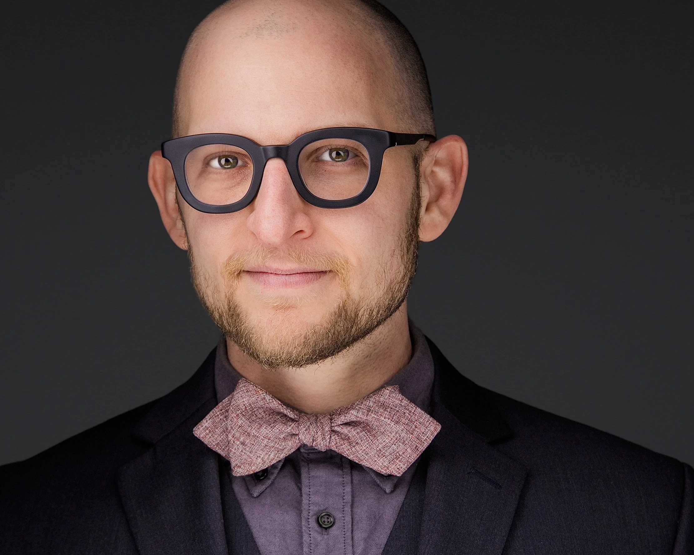 A man with glasses and a beard wearing a dark suit, gray shirt, and pink bow tie, posing against a dark gray background.
