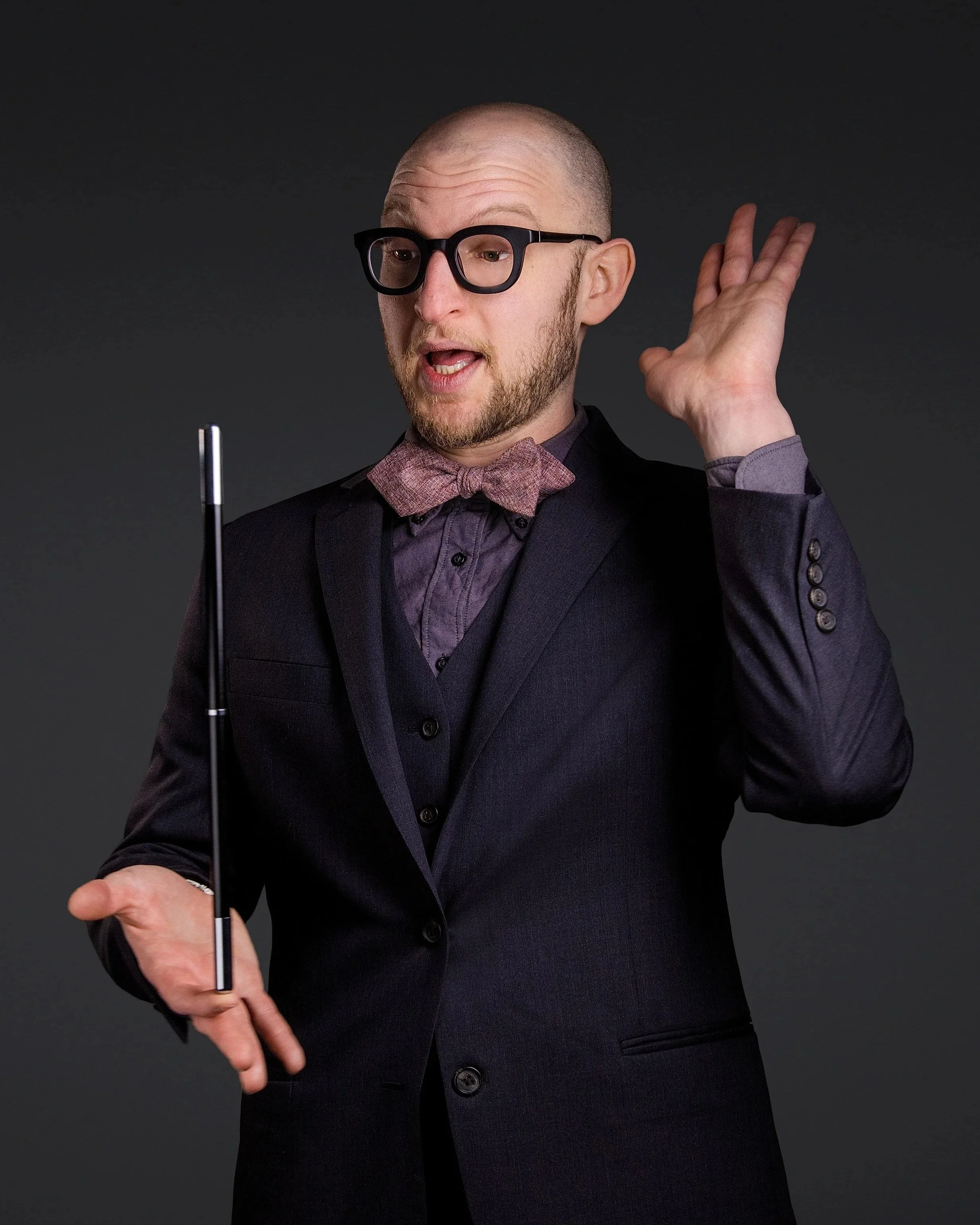 A man with glasses, a beard, and a shaved head, wearing a dark suit with a purple shirt and bow tie, holding a conductor's baton and gesturing with his other hand against a dark background.