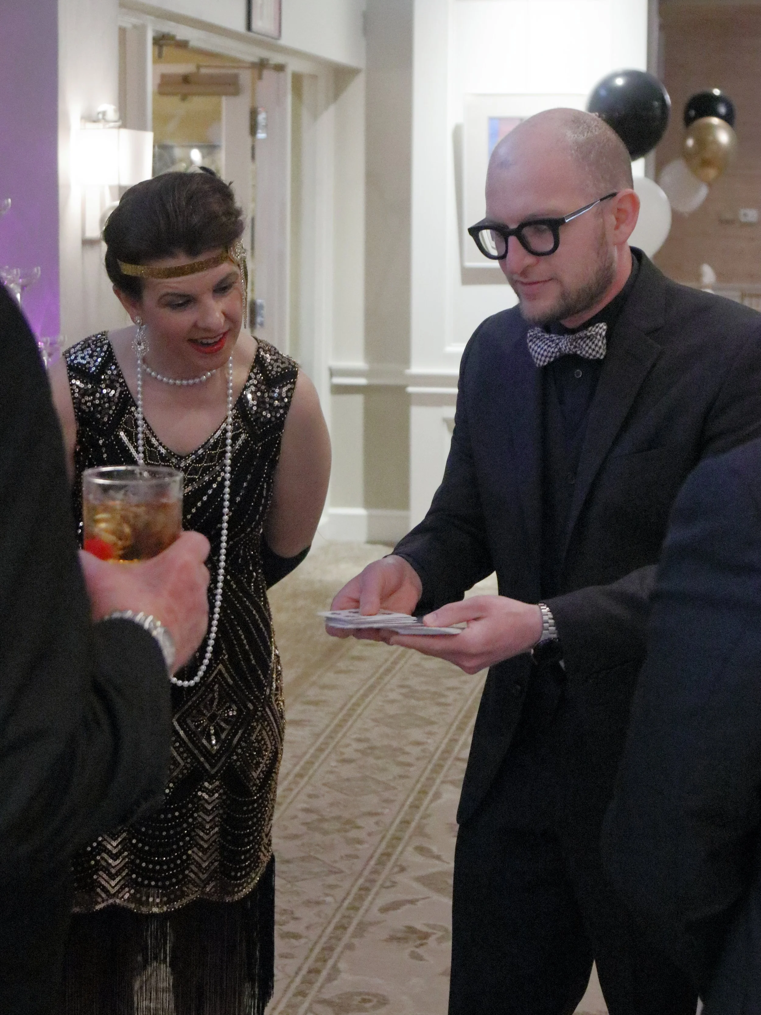 A woman in a vintage 1920s style dress with beaded details and pearl accessories is amazed by a card trick performed by a man wearing a suit with glasses and a bow tie at a social event, with balloons and decor in the background.