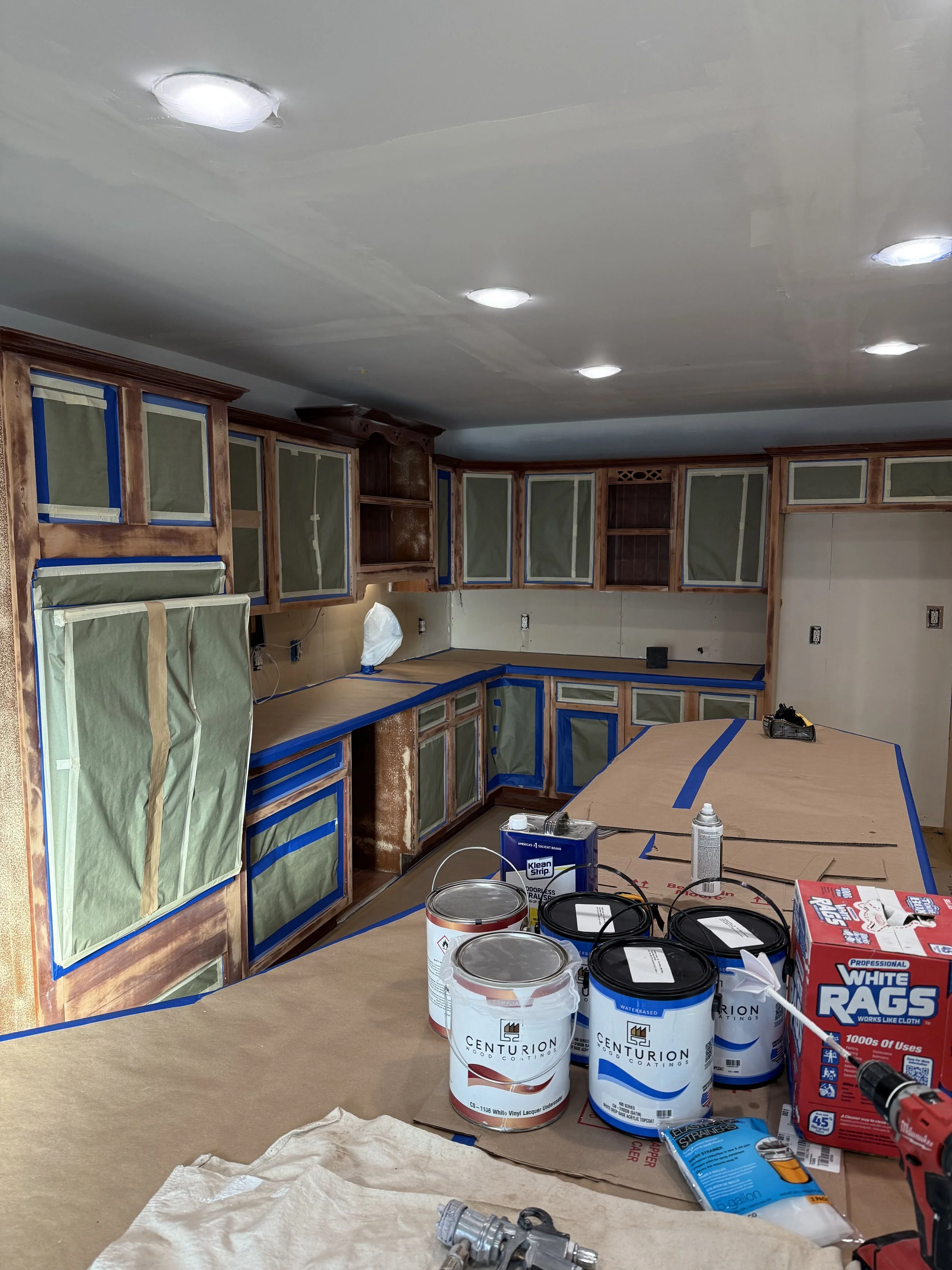 Kitchen under renovation with cabinets covered in painter's paper, various cans of paint, a spray can, a box of white rags, and tools on the counter.