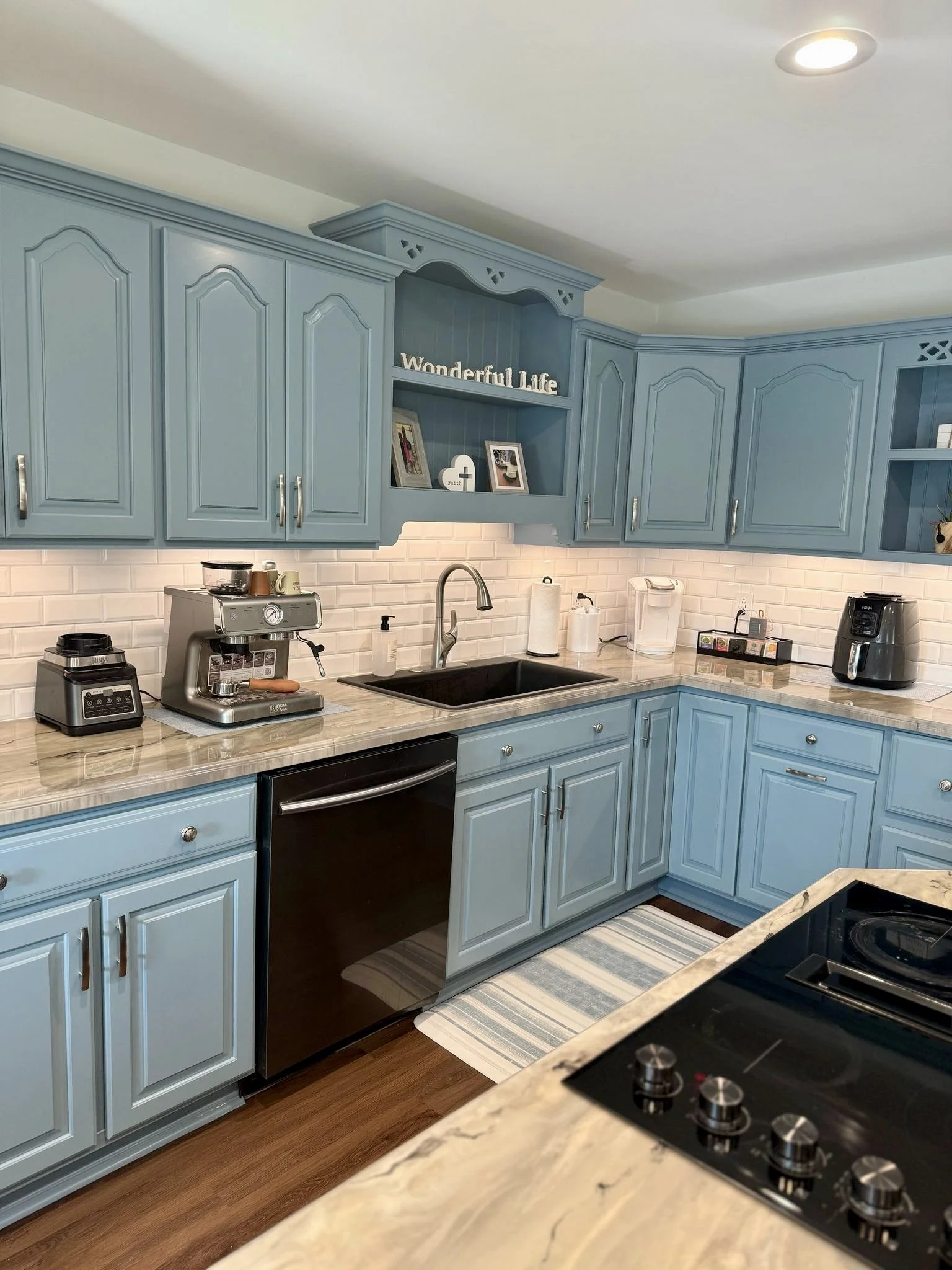 Blue kitchen cabinets with granite countertops, a black sink, and various appliances including an espresso machine, blender, and air fryer. Decor includes a sign reading 'Wonderful Life' and framed photos.