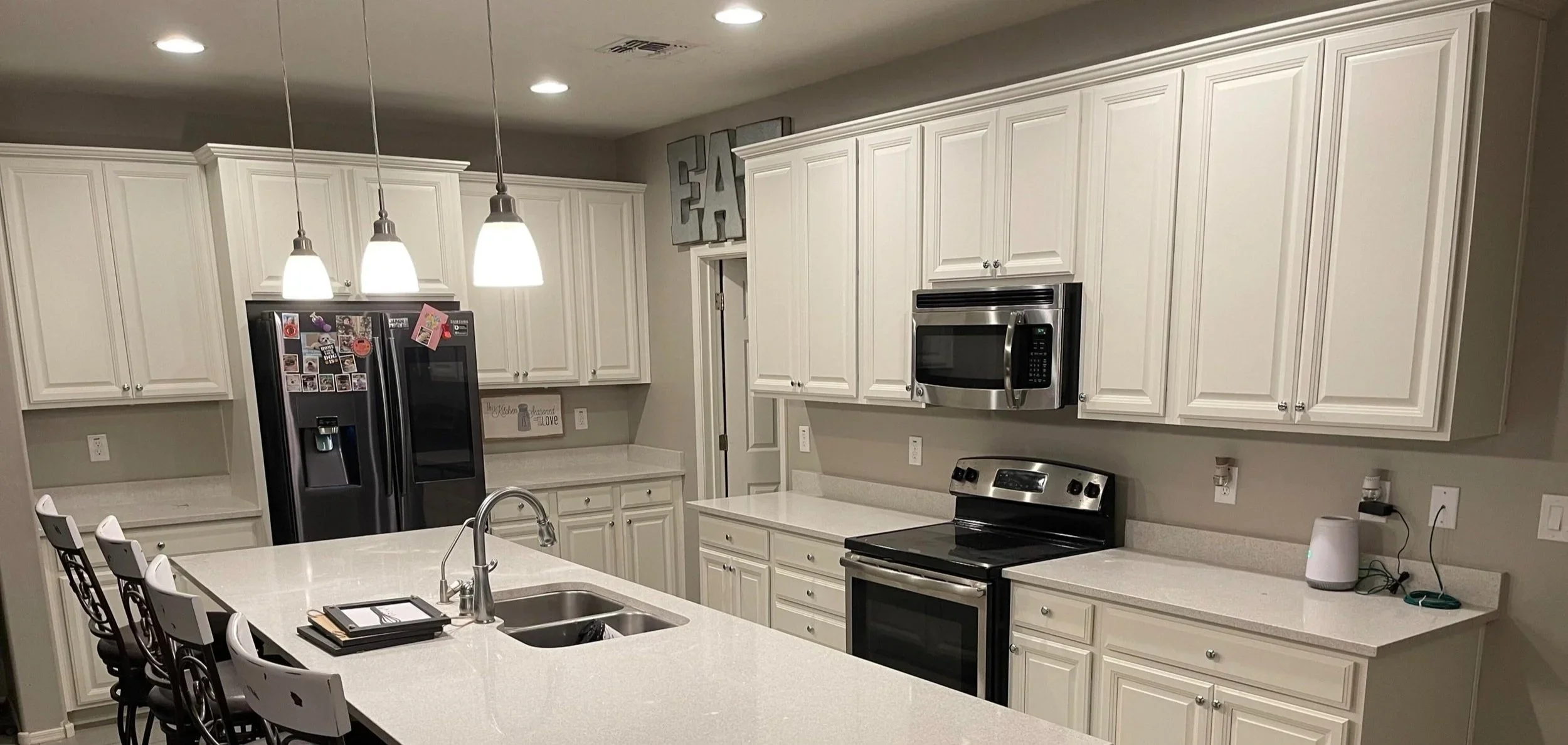 Modern kitchen with white cabinets, stainless steel appliances, a black refrigerator, microwave, oven, and a white island with a sink. Overhead lighting and barstools along one side of the island.