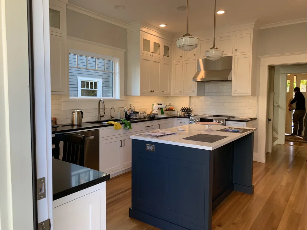 A modern kitchen with white cabinets, a navy blue island, a black countertop, stainless steel appliances, and hardwood floors. There are two pendant lights above the island, and a window above the sink.