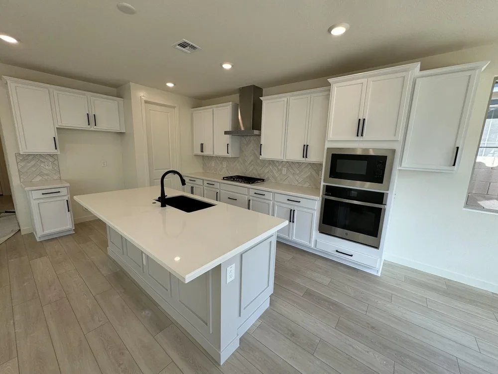 Modern kitchen with white cabinetry, black hardware, a kitchen island with a black sink and faucet, built-in oven and microwave, stainless steel range hood, light wood flooring, and a window.