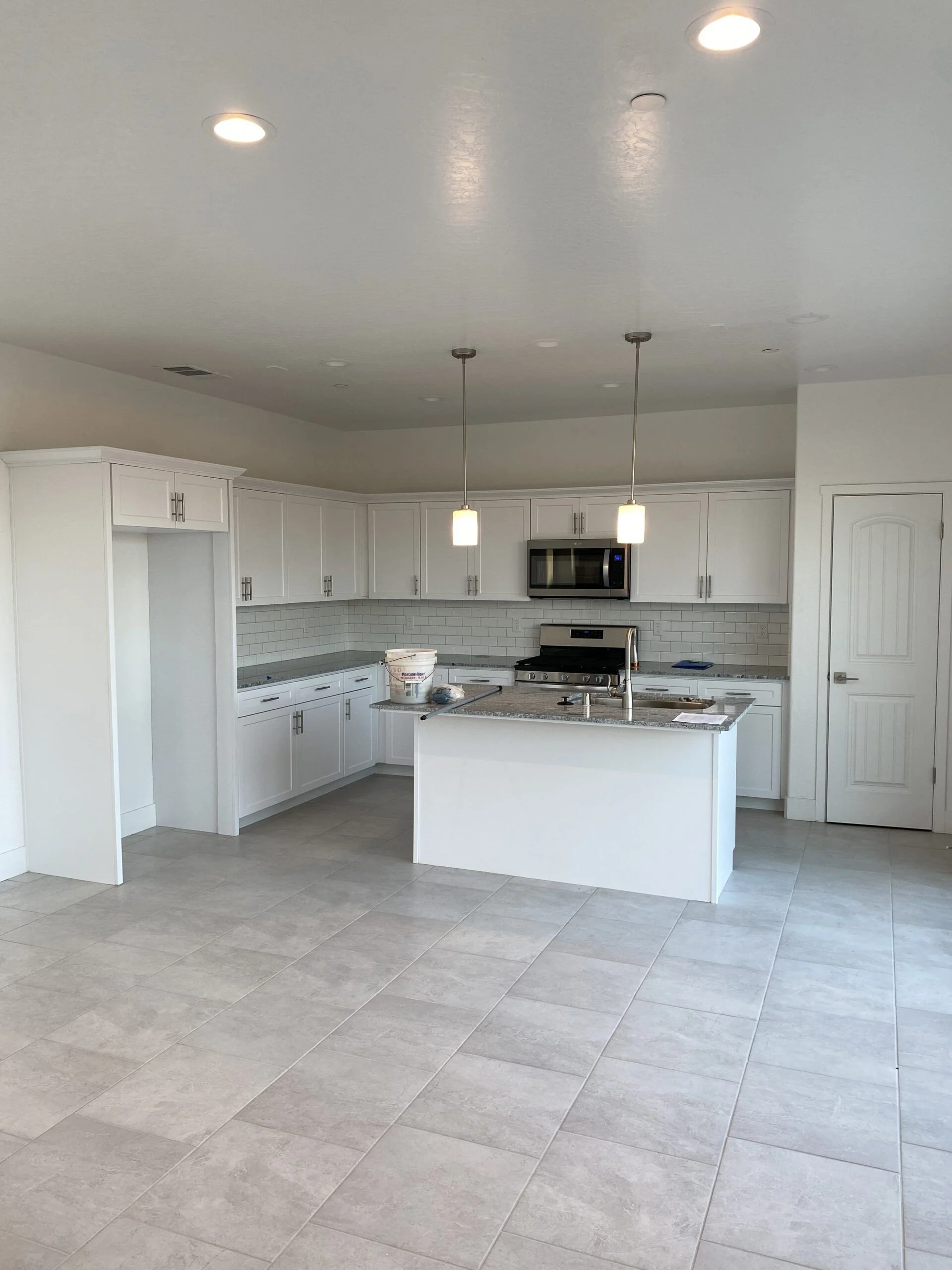 A modern white kitchen with white cabinets, stainless steel appliances, and a kitchen island with a granite countertop. There are two pendant lights above the island, and the kitchen features a tile backsplash and beige floor tiles.