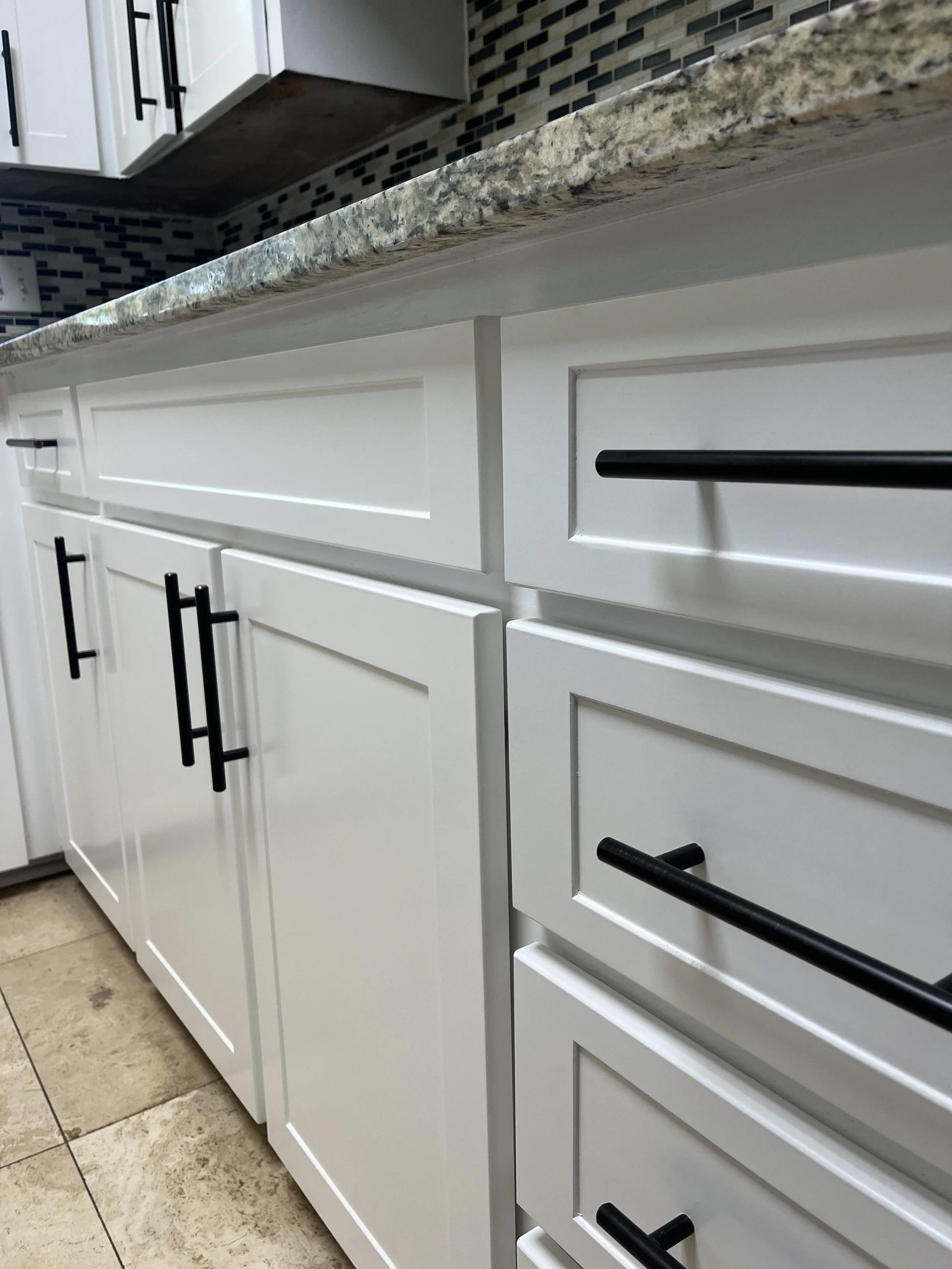 Close-up of white kitchen cabinets with black handles and granite countertop, tiled backsplash, and part of the kitchen floor.