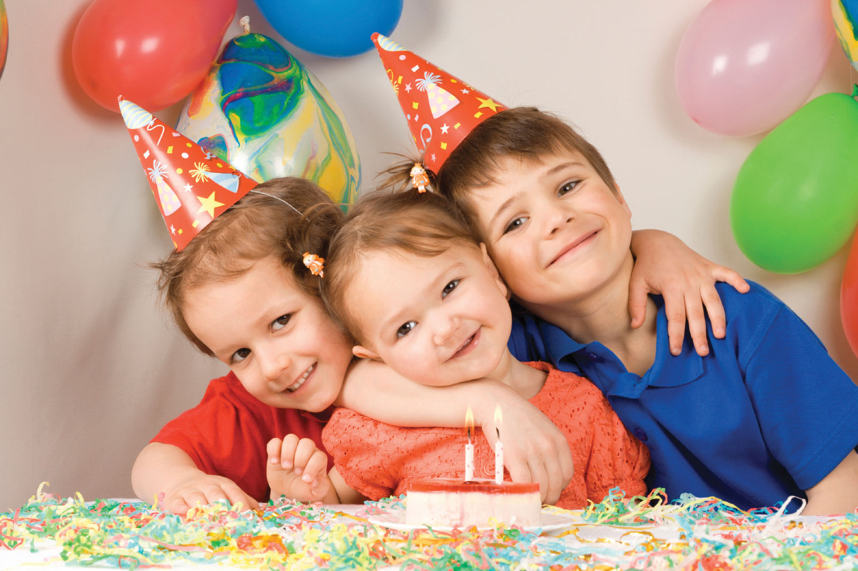 Three children celebrating a birthday with party hats, colorful balloons, a cake with lit candles, and confetti.