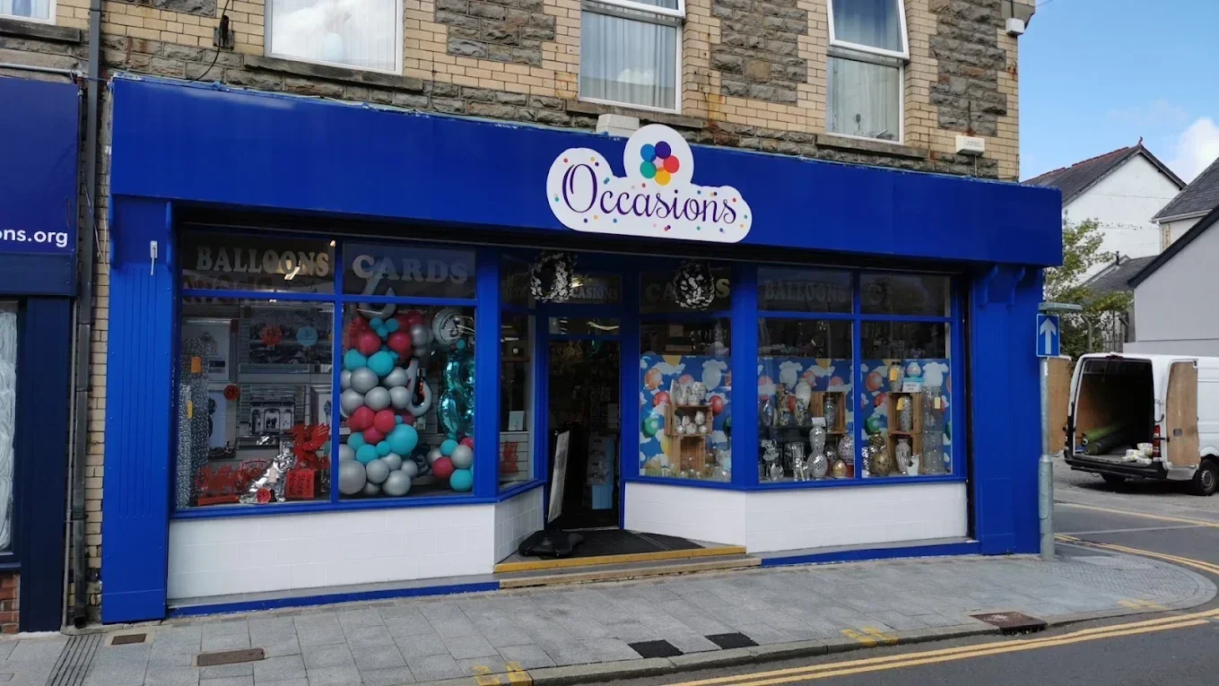 A storefront with a blue exterior and large windows displaying balloons and decorations. The store sign reads 'Occasions' with colorful dots above it. There are balloons arranged in a column inside the window and various party supplies visible.