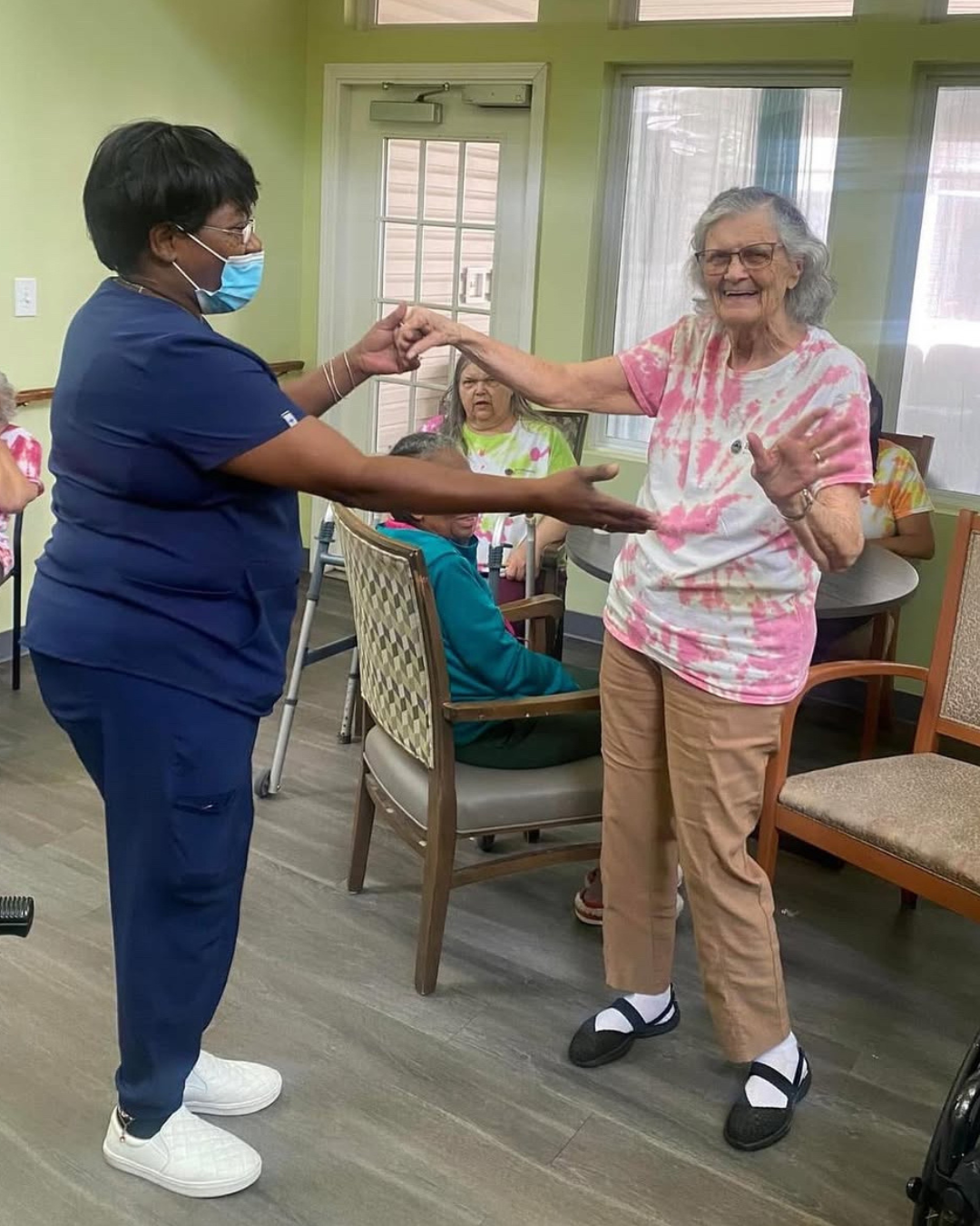 A healthcare worker and an elderly woman are dancing together in a room with other seated elderly individuals, some wearing tie-dye shirts, and a green wall in the background.