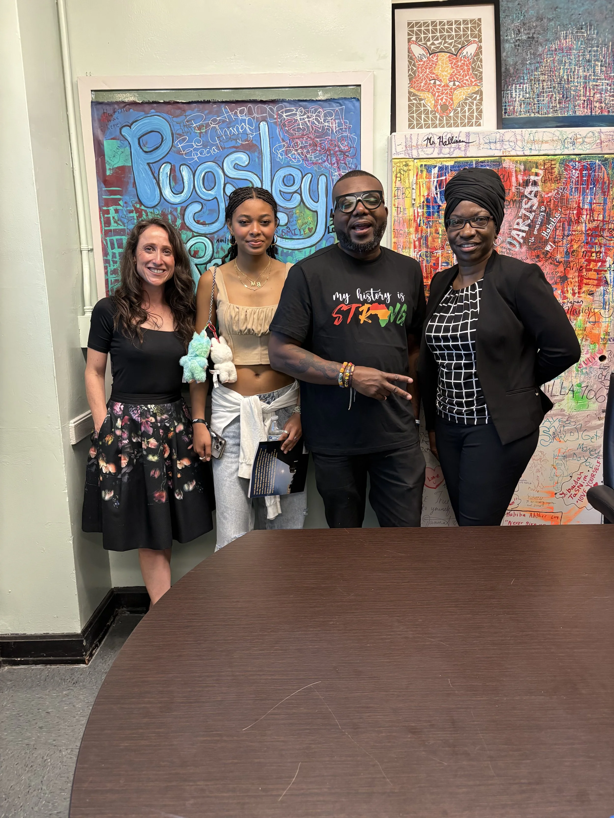 Four people stand together in front of colorful artwork, posing for a photo in an indoor setting. They are smiling and dressed casually, with one person wearing glasses and another wearing a black headwrap.