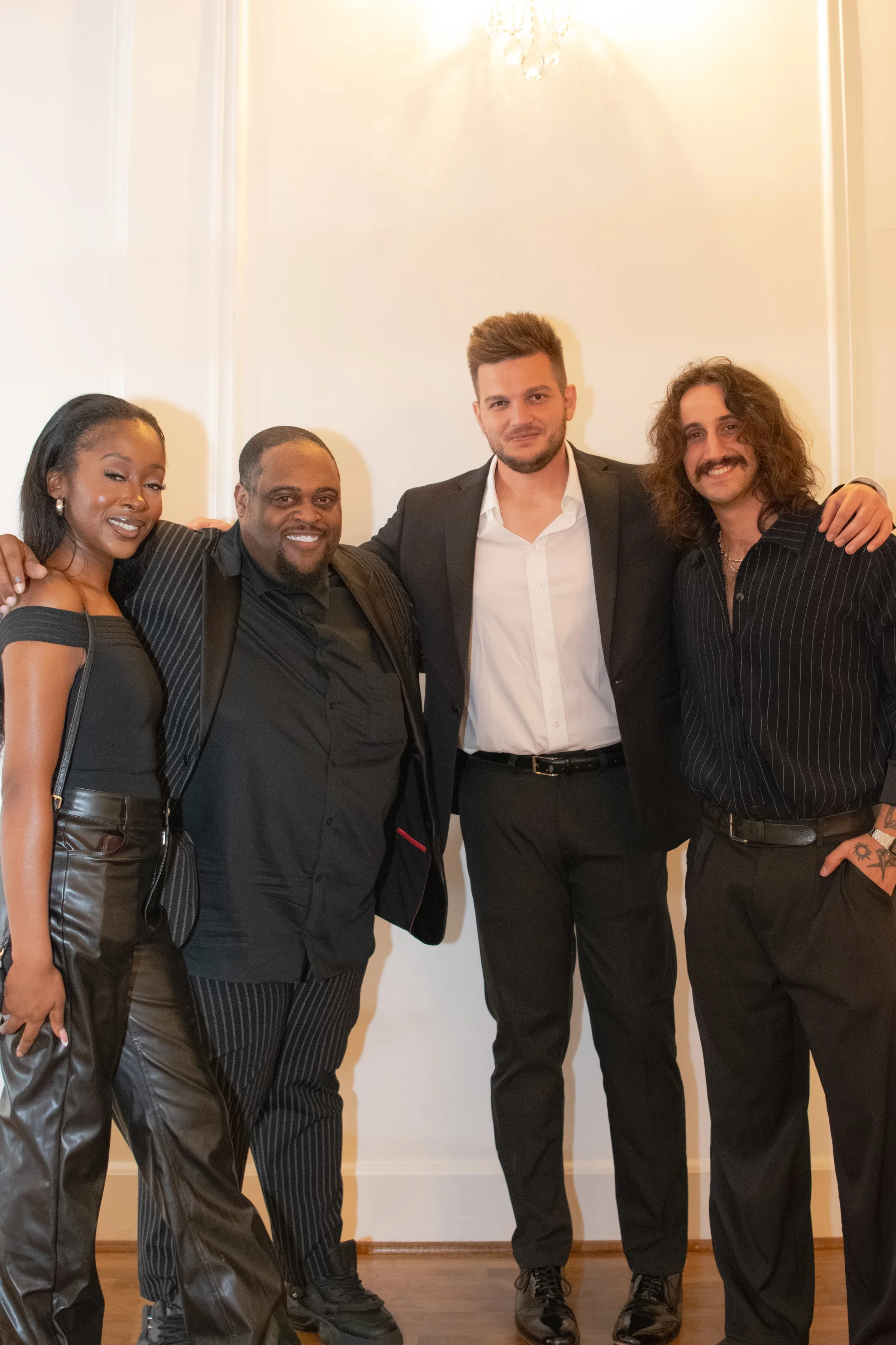 Group of four people in formal attire standing together indoors with a plain wall background.