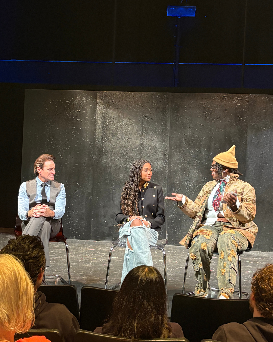 Three people seated on stage during a panel discussion, with audience members visible in the foreground, set against a dark backdrop.