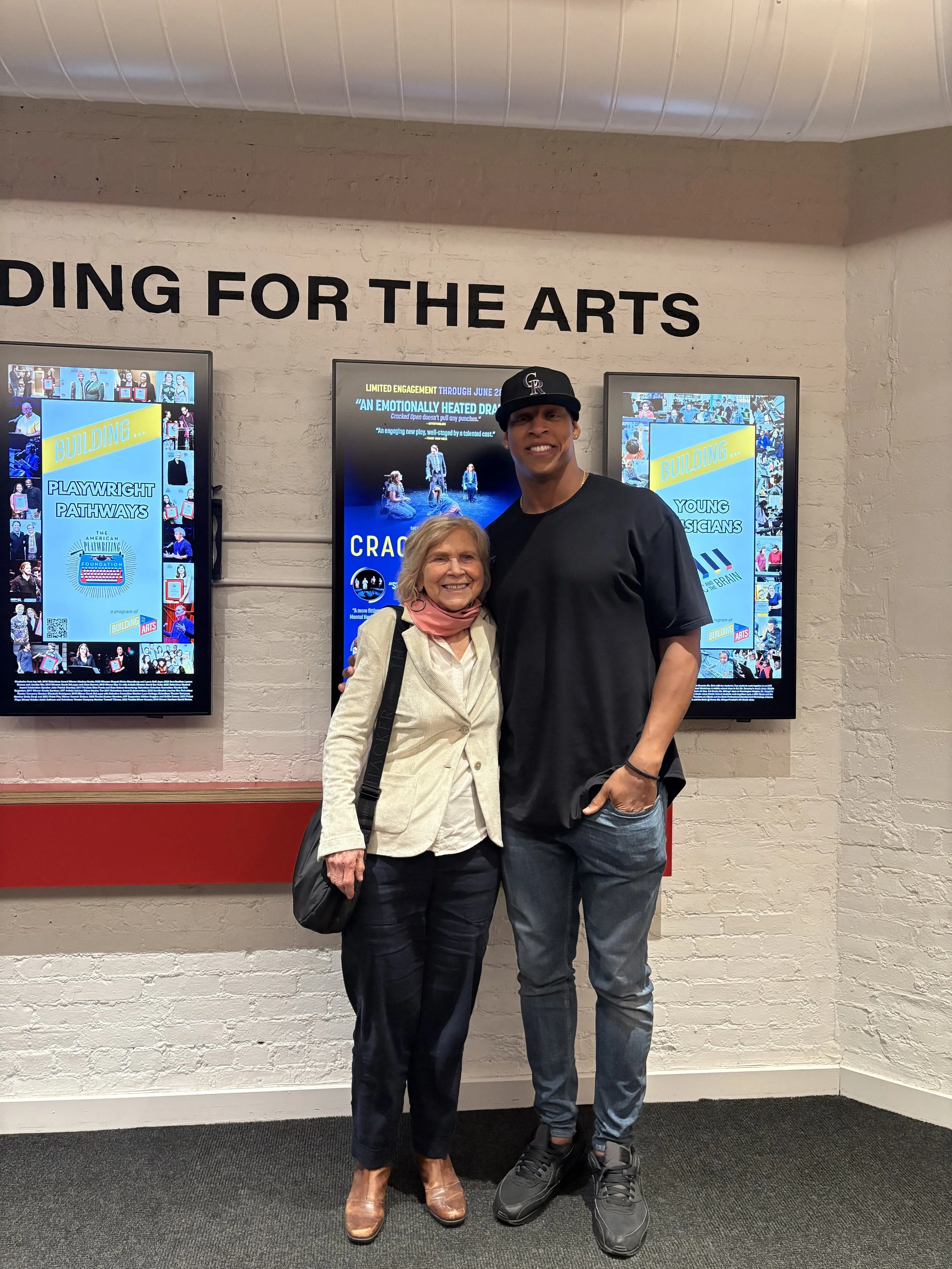 A woman and NFL veteran Jack Crawford standing together, smiling, in front of posters advertising a theatrical production called Cracked Open in New York on Theater Row.