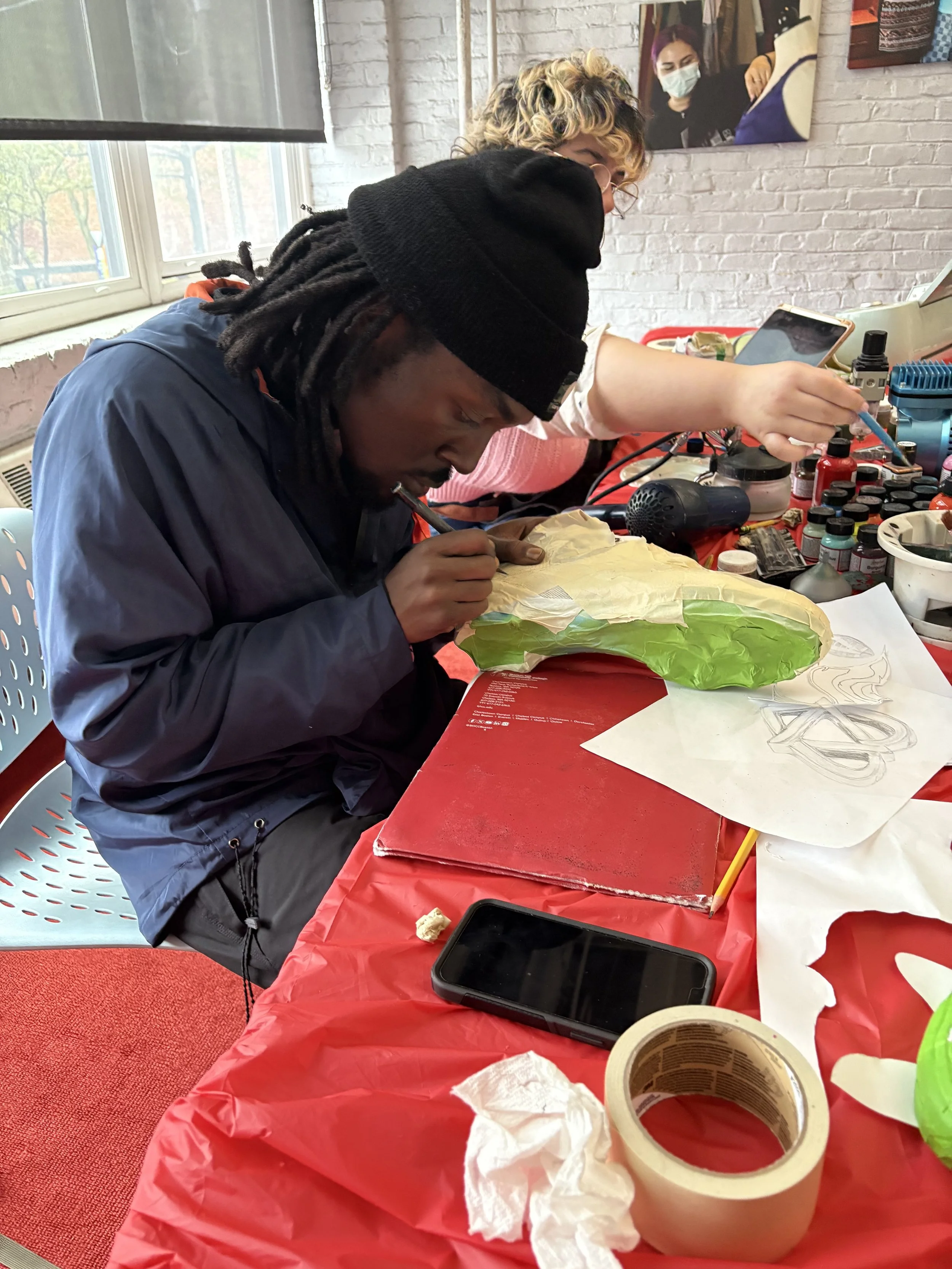 Two people working on arts and crafts at a table covered with a red tablecloth, surrounded by art supplies, in a room with a brick wall and a window.