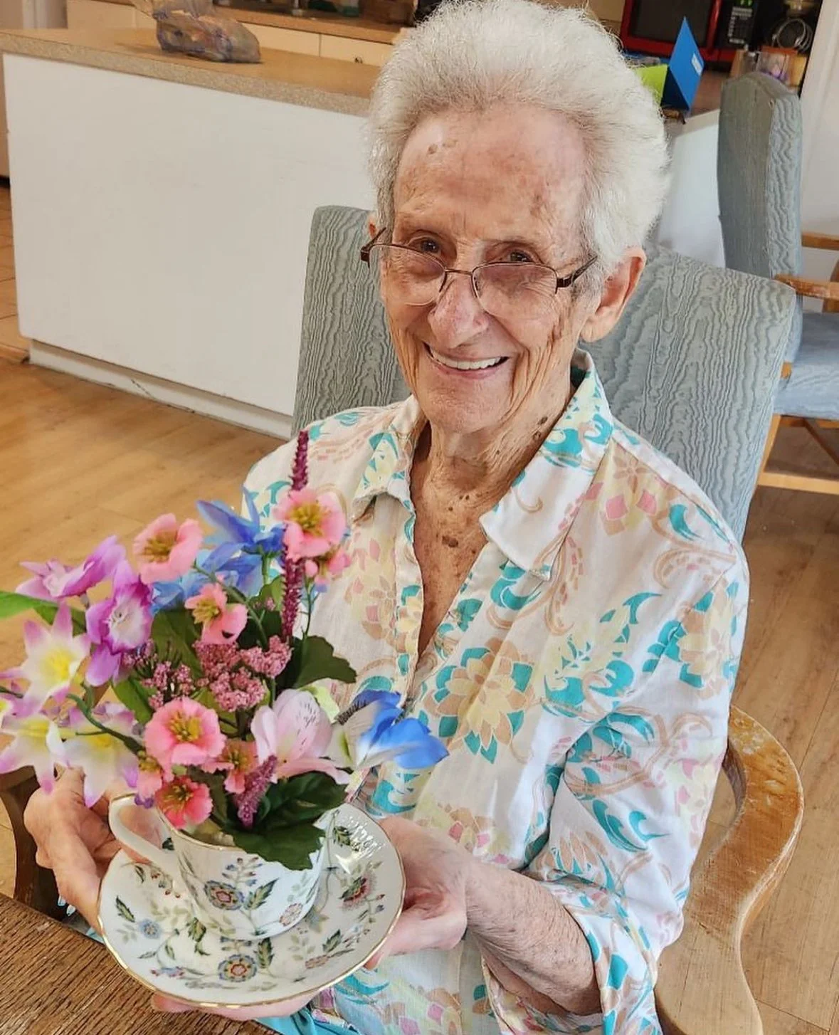 An elderly woman with gray hair, glasses, and a floral shirt, smiling as she holds a teacup filled with pink and purple flowers, sitting in a light-colored armchair in a cozy home setting.
