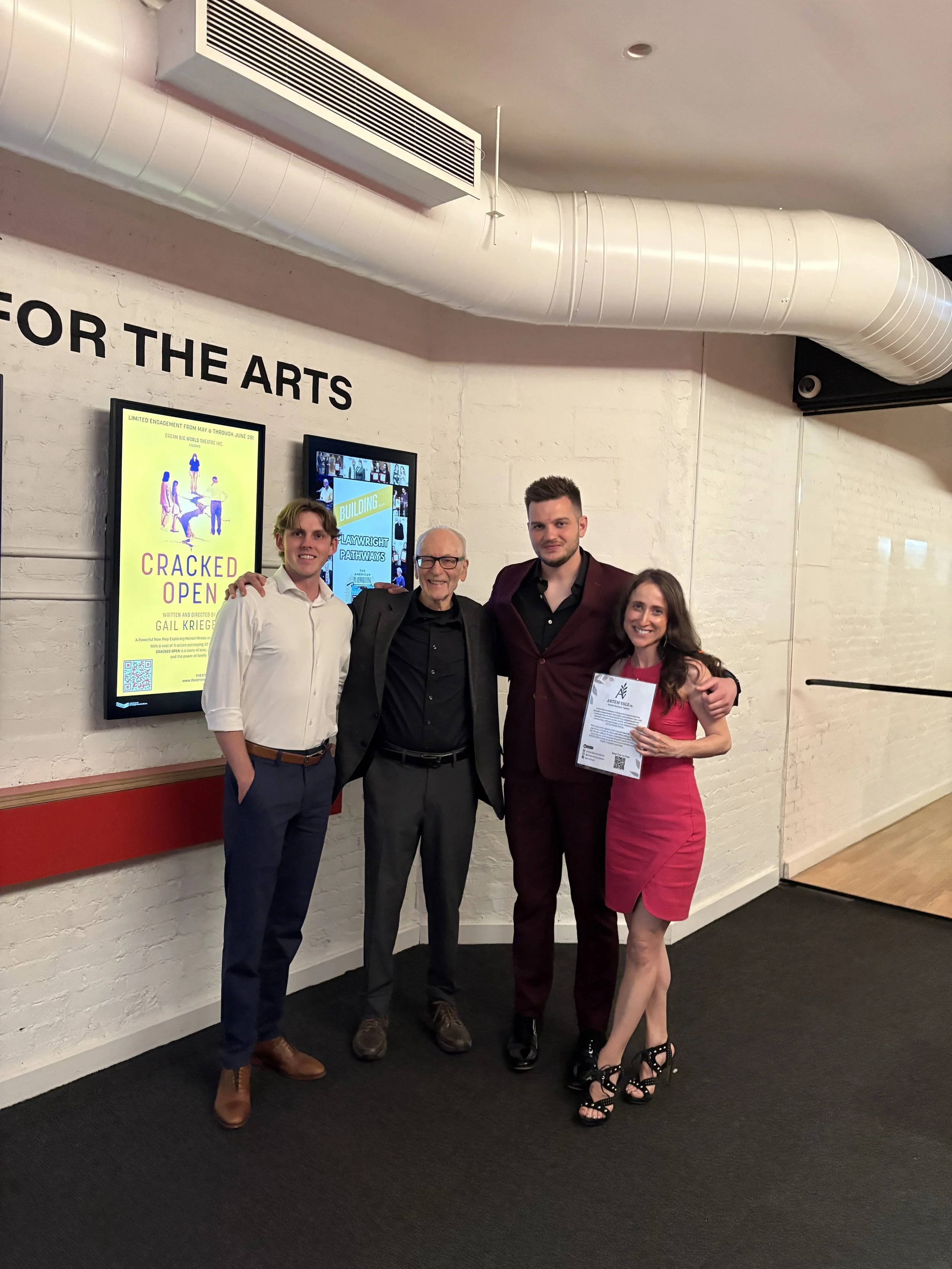 Hayden grove and individuals standing together in a hallway, smiling. The person on the right is holding a certificate. There are digital screens and posters on the wall behind them, including one for a play called 'Cracked Open.'
