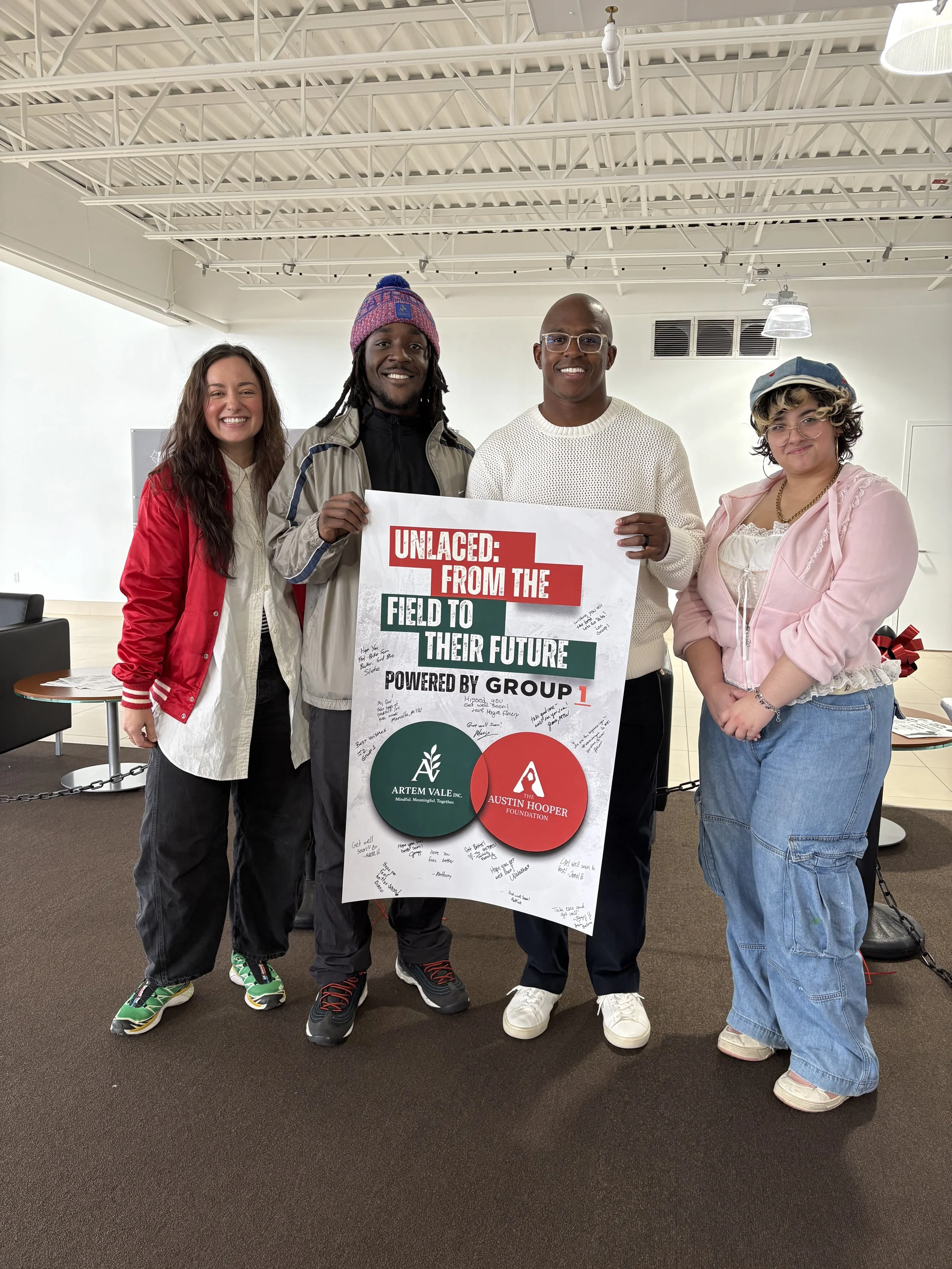 Four people standing indoors holding a large sign that reads 'Unlaced: From the Field to Their Future Powered by Group 1,' with signatures and messages written around it. The people are smiling and dressed casually, with a plain white wall and ceiling in the background.
