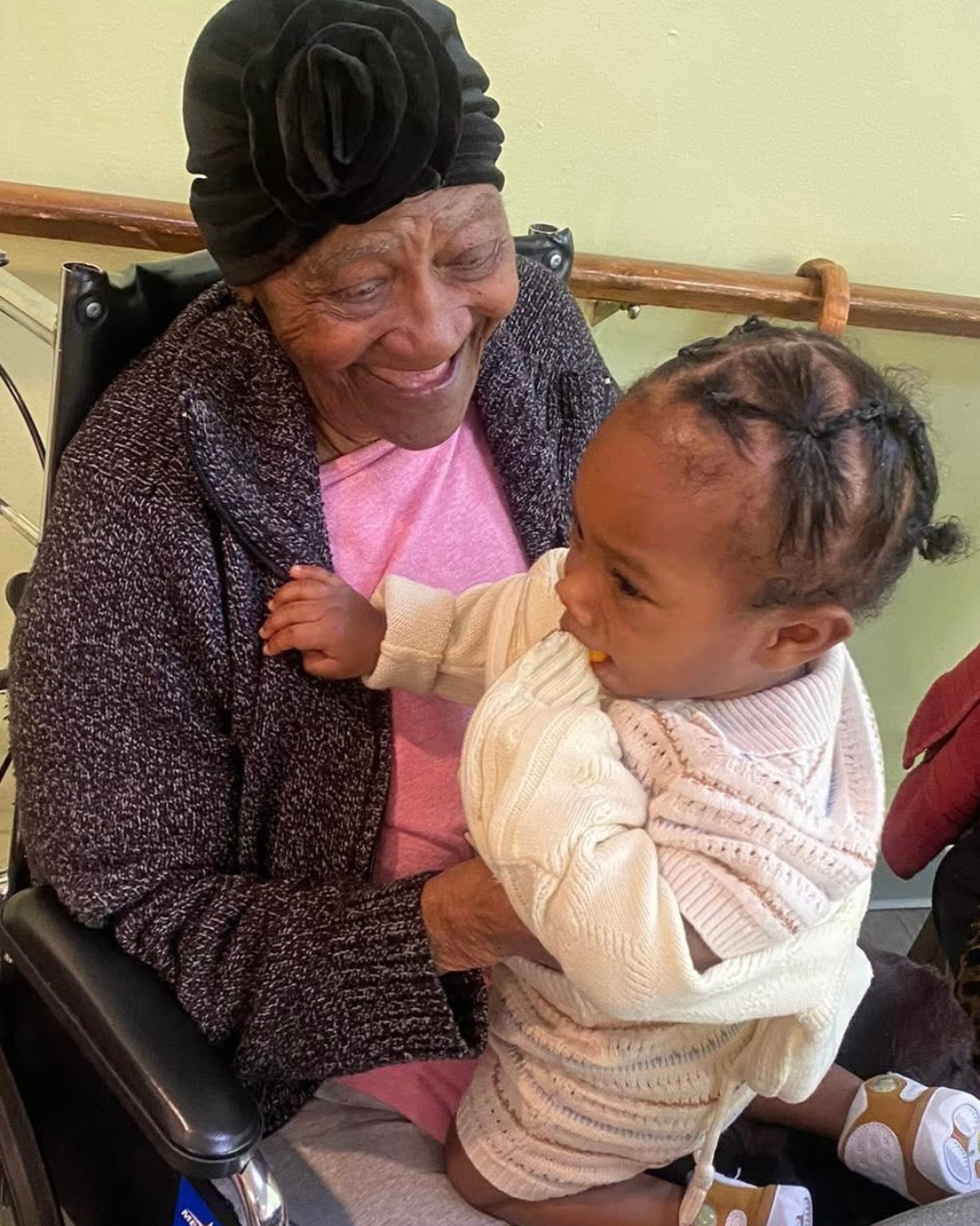 An elderly woman in a wheelchair smiling as she holds a young girl, who is sitting on her lap and looking at her, in a warm embrace indoors.