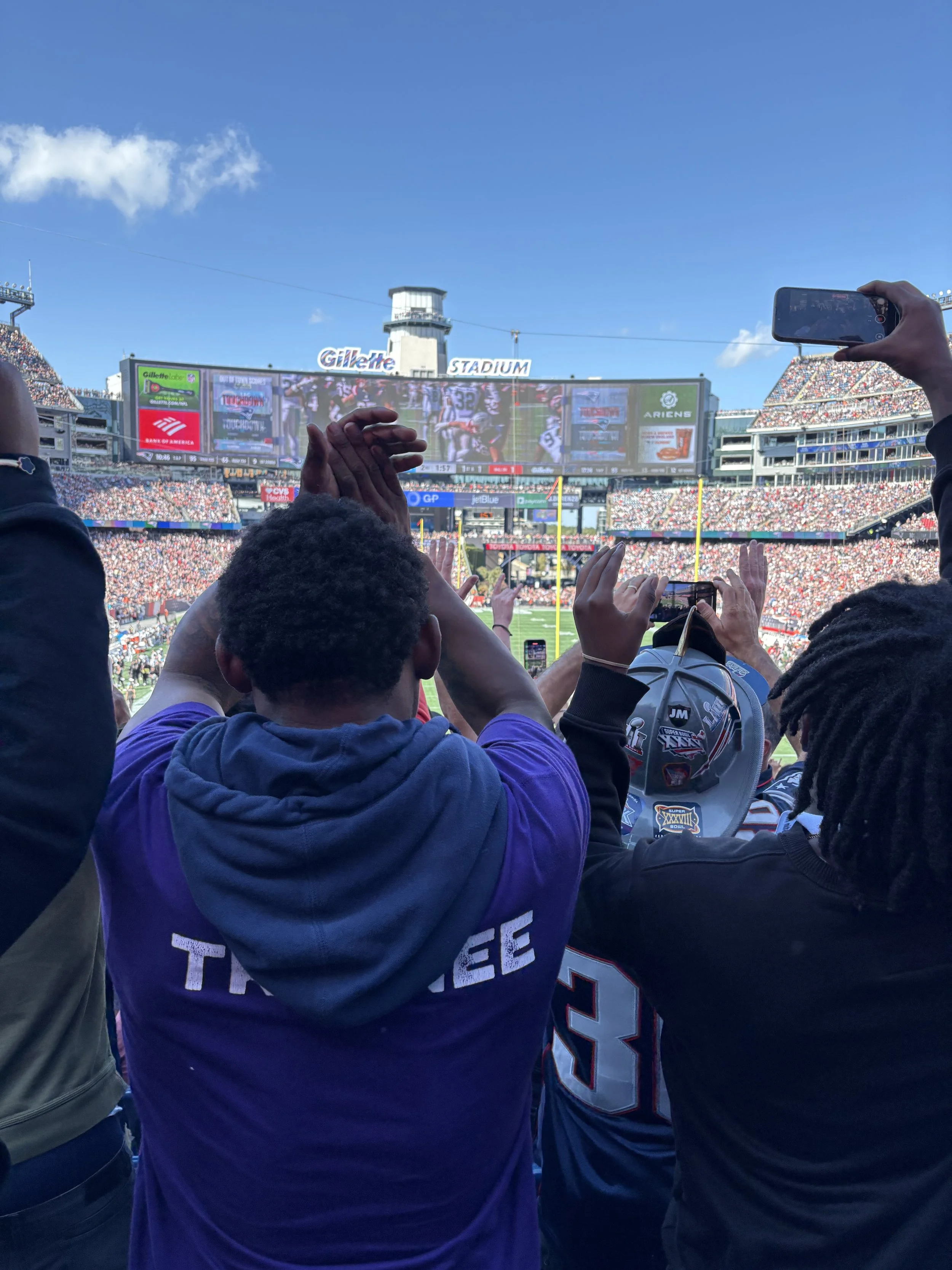 View of fans at a football game in a stadium with the field and scoreboard visible in the background, crowd seated in the stands, and some fans taking photos or videos with their phones.