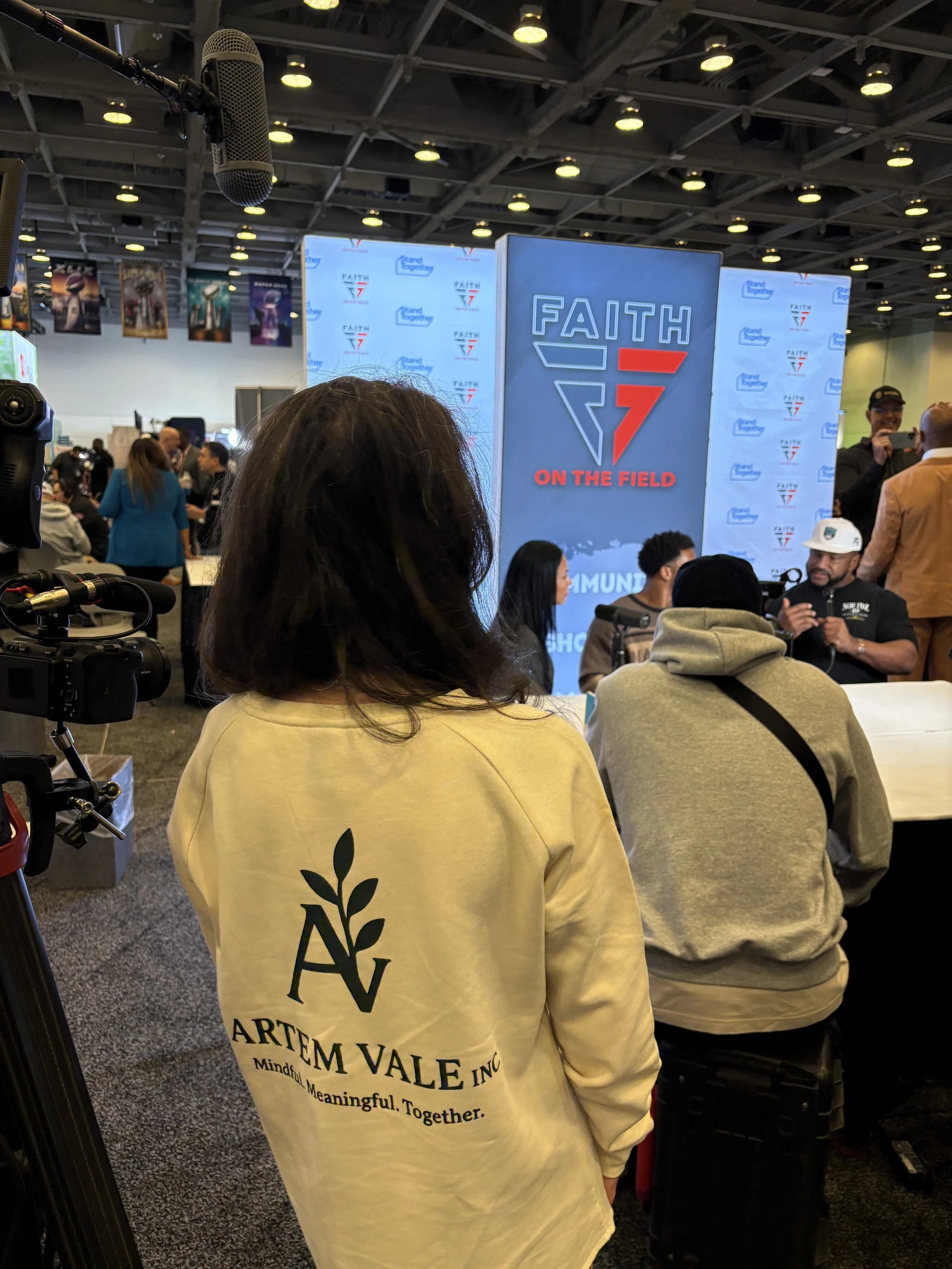 A woman with long dark hair wearing a cream-colored sweatshirt with the Artem Vale Inc logo and slogan, standing indoors at a conference or event. In the background, there are multiple people, some sitting and some standing near a large digital screen displaying the logo for 'Faith on the Field', with a stage and various banners overhead.