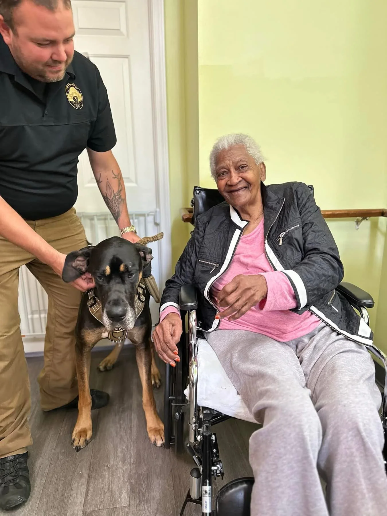 An elderly woman in a wheelchair smiling at the camera with a man and a large dog beside her. The man is holding the dog, and the woman is wearing a black jacket, pink shirt, and gray sweatpants. The background shows a light green wall and a white door.