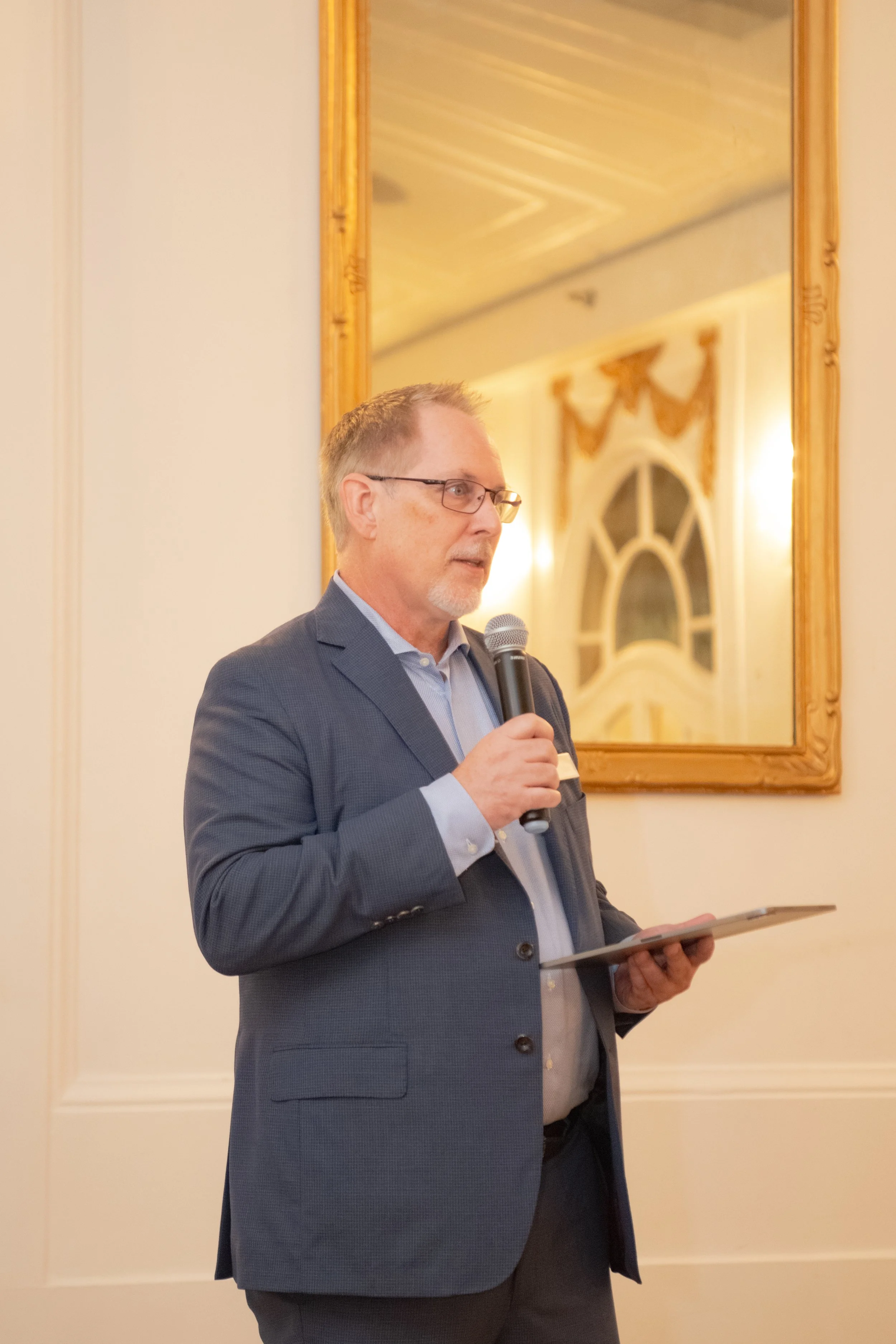 A middle-aged man in a blue suit, glasses, and a light blue shirt, holding a microphone and a tablet, speaking at a formal event in a room with elegant decor, including a large mirror and ornate window treatments.
