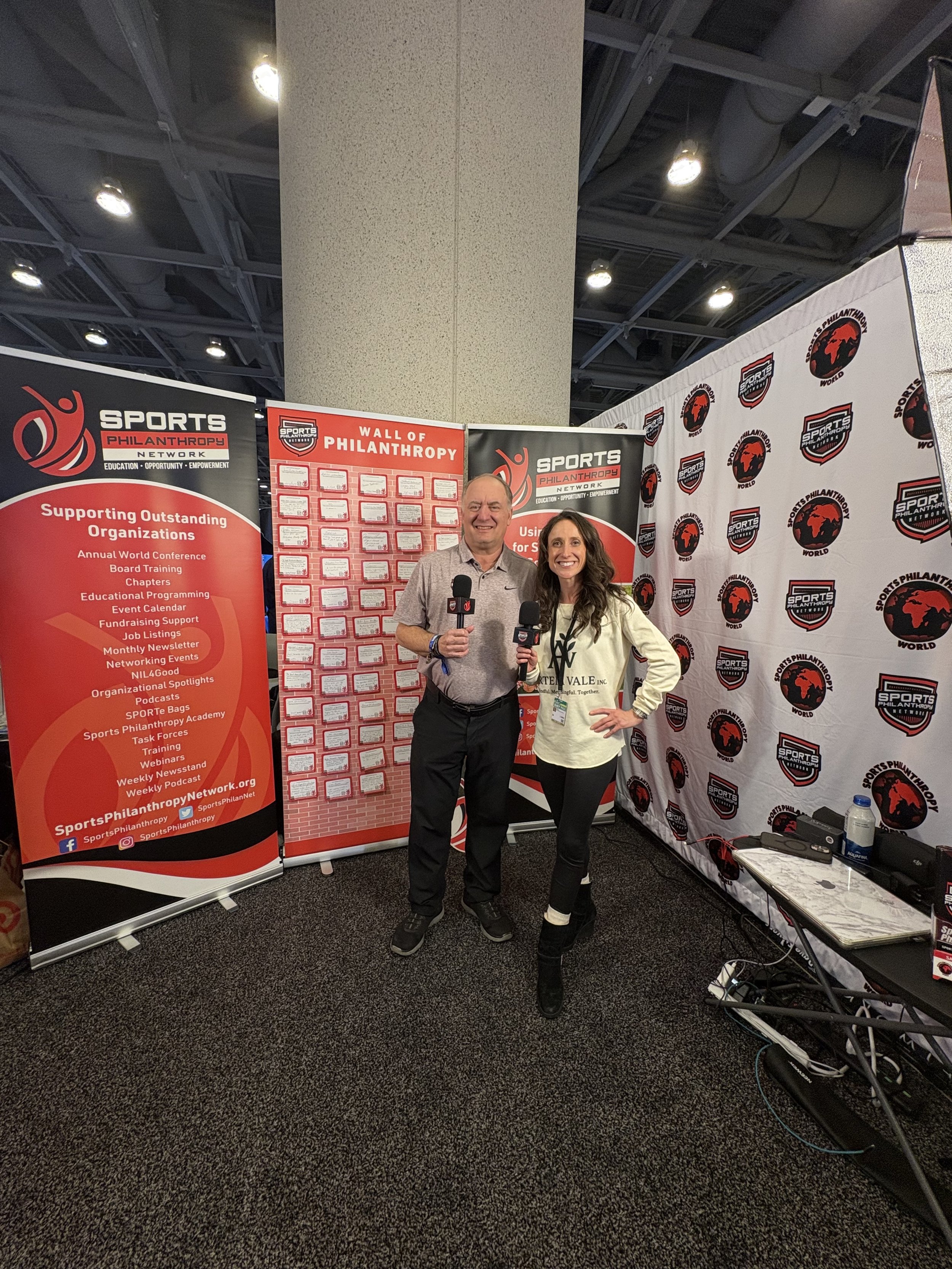 Arian May with Sports Philanthropy Network standing in front of sports philanthropy banners at an indoor event, holding microphones, smiling for the camera.