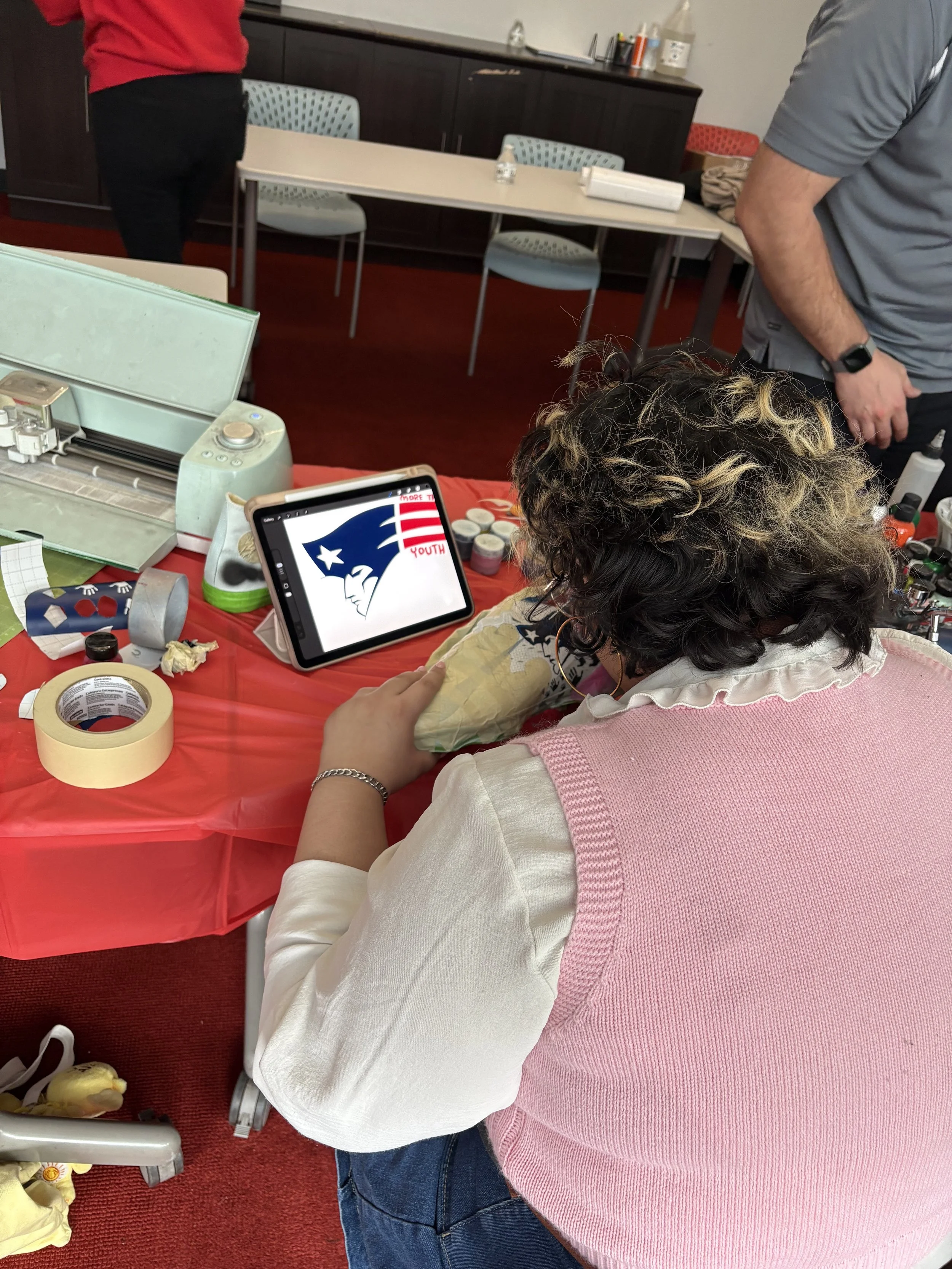 A woman is sitting at a table with crafting supplies, looking at a tablet displaying a New England Patriots logo. She is surrounded by tape, paper, and other crafting materials, in an indoor setting with several empty chairs and a red carpeted floor.