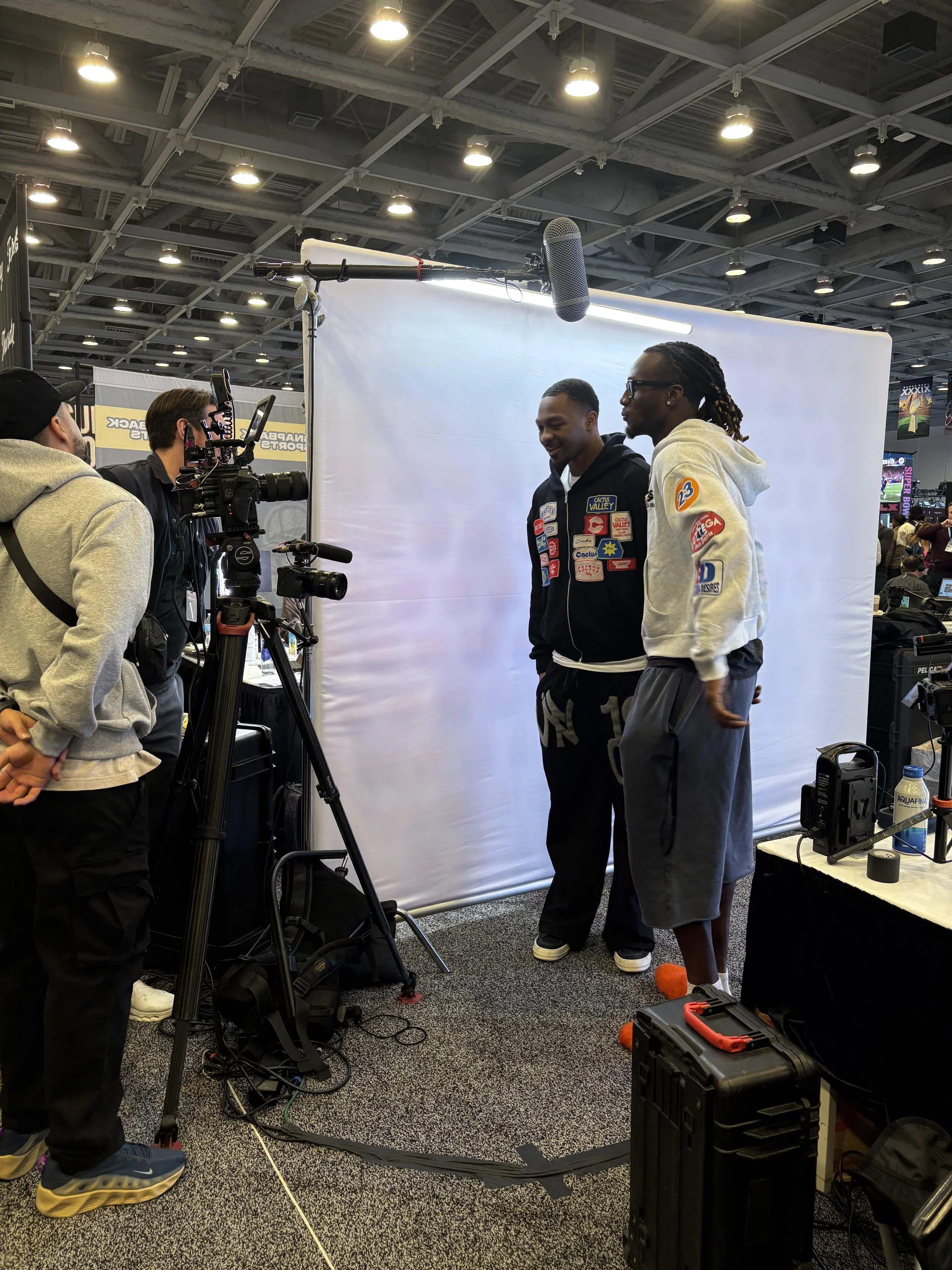 Two men are being interviewed on camera at a convention. They are standing in front of a white backdrop, with one man smiling and the other talking. Several people and equipment, including cameras, microphones, and case boxes, surround them in a large, indoor venue with a high ceiling and bright lights.