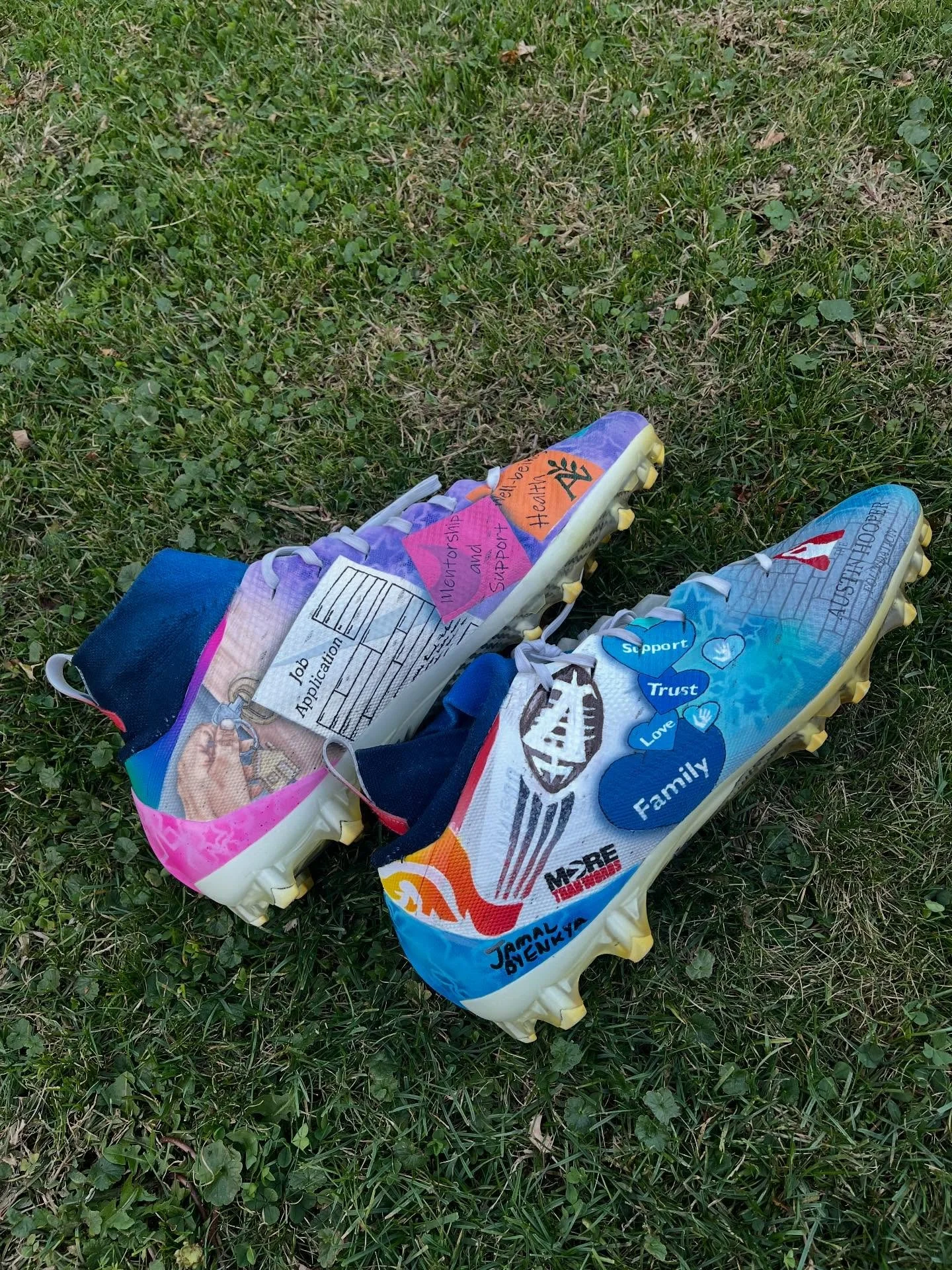 Colorful soccer cleats decorated with various stickers, drawings, and messages, lying on green grass.