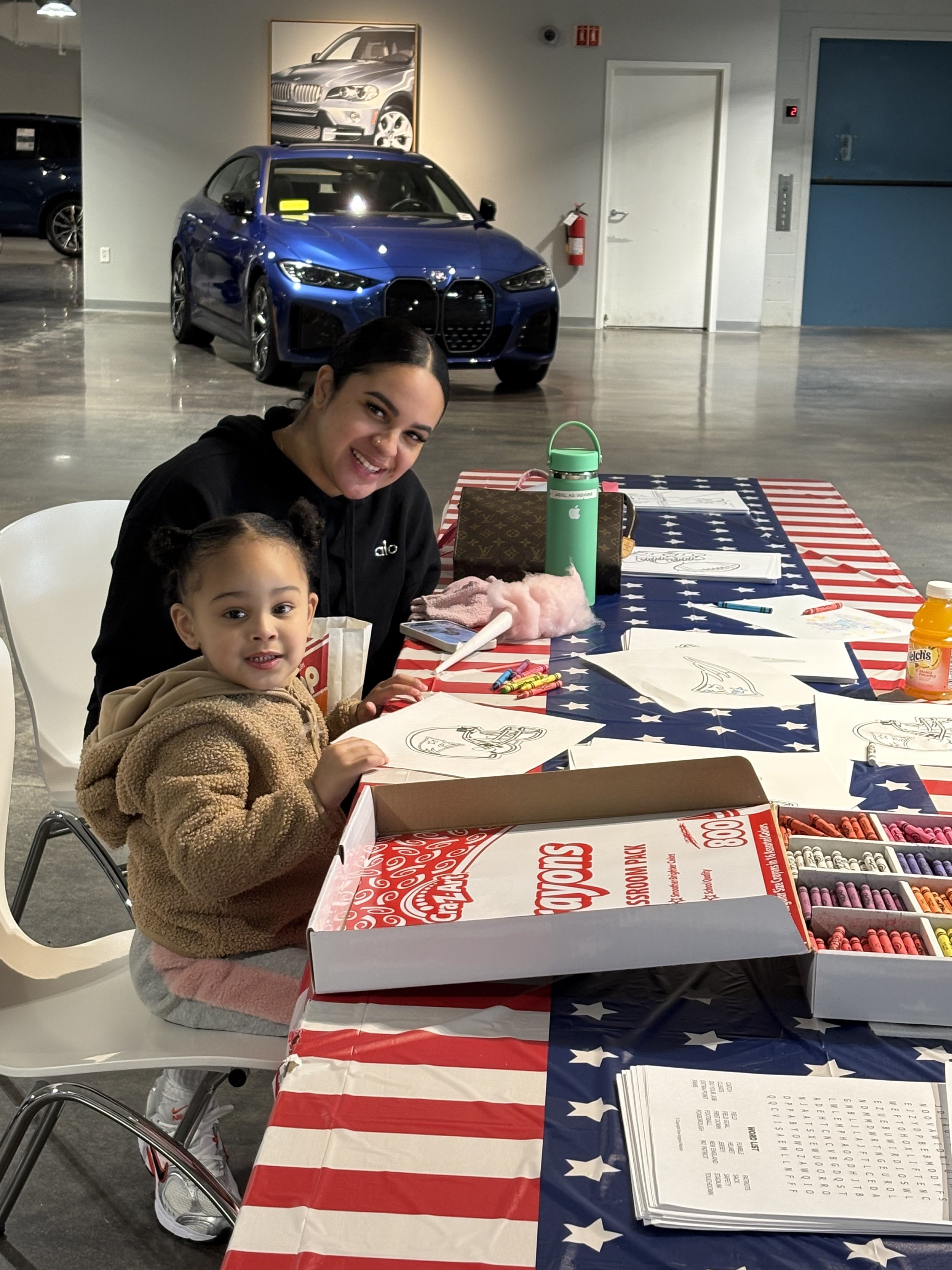 A smiling woman and young girl sitting at a table decorated with an American flag-themed tablecloth, engaging in coloring activities with printed sheets and crayons. In the background, a blue car is parked inside a showroom or garage, with a framed car poster on the wall.