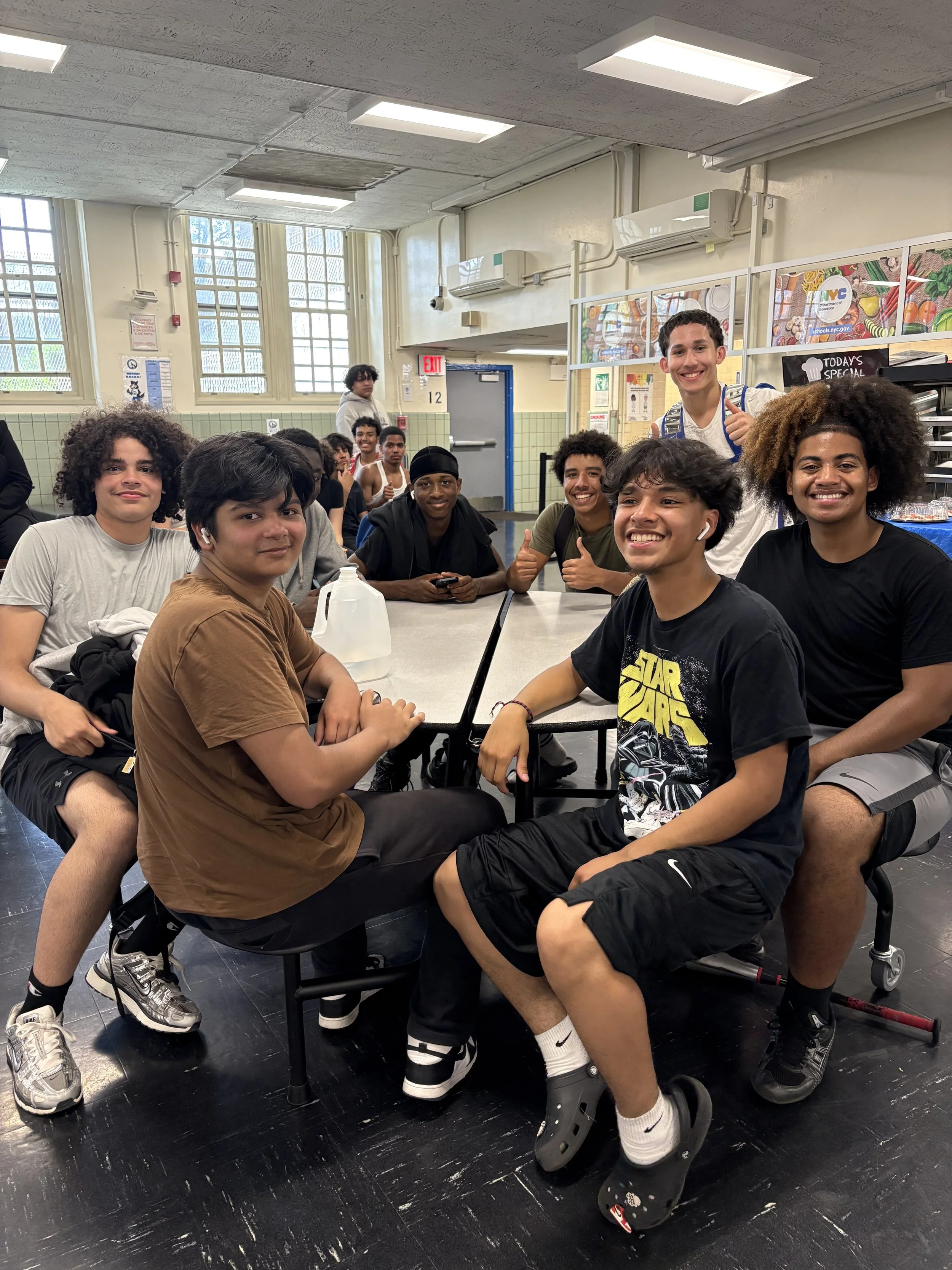 A group of young boys sitting and standing around a school lunch table in a cafeteria, smiling and giving thumbs up.