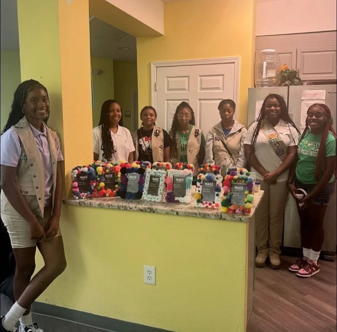 Group of women standing behind a decorated table with framed crafts, in a kitchen area.