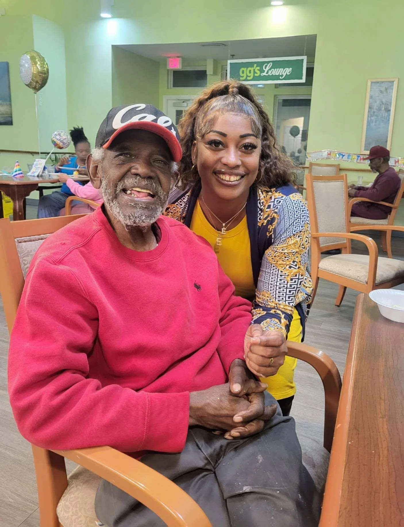 Smiling elderly man with a grey beard, wearing a red sweatshirt and a cap, sitting in a chair and holding hands with a young woman in a yellow shirt and patterned jacket, both smiling in a lounge area decorated for a celebration.