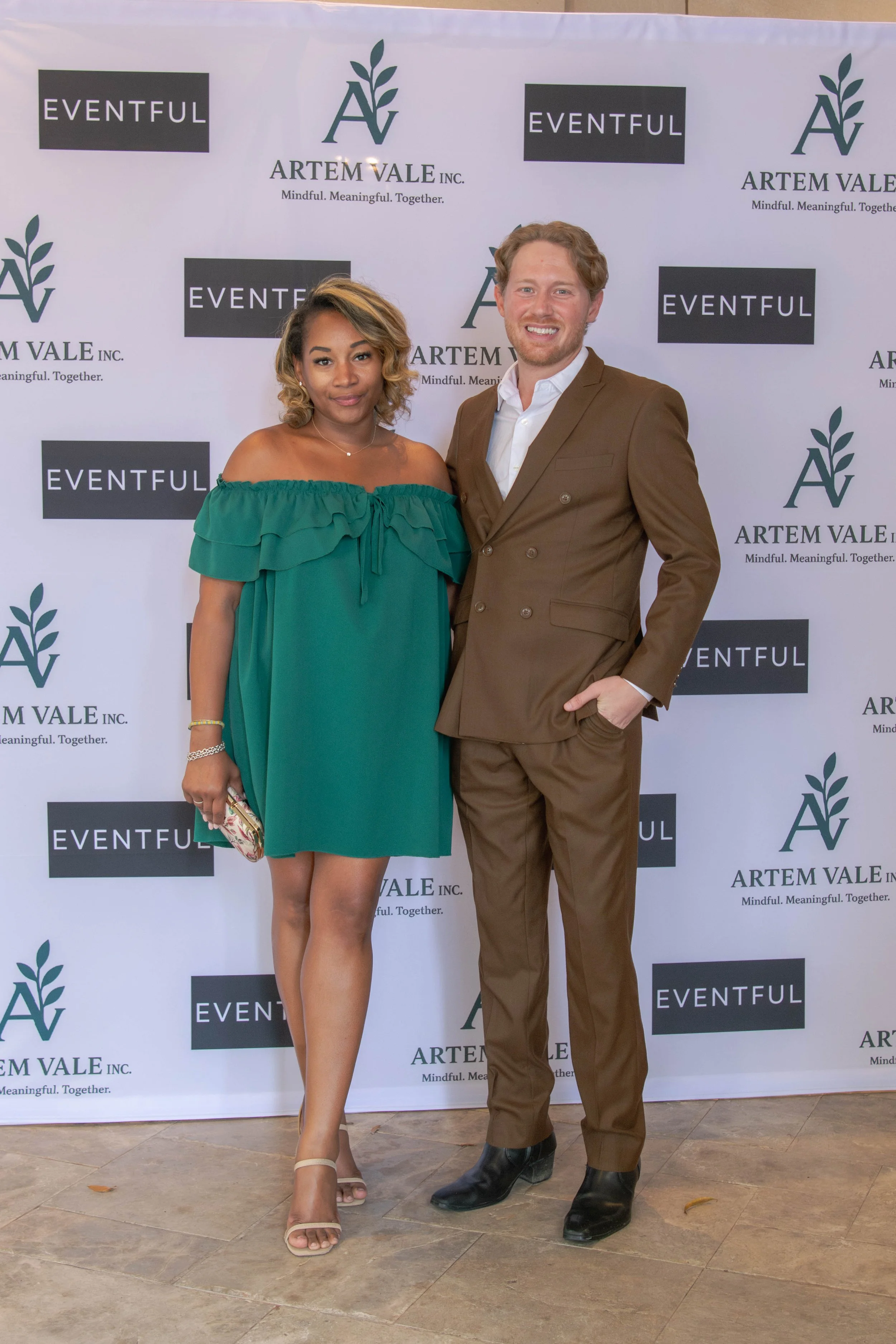 A woman and a man standing in front of a backdrop with logos for Artem Vale Inc. and Eventful. The woman is wearing an off-the-shoulder green dress and holding a small clutch. The man is wearing a brown suit and black shoes. They are smiling and posing for the photo.