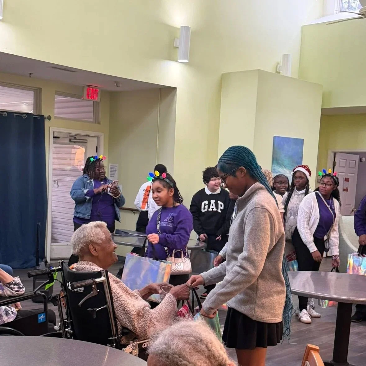 Young woman giving a gift to an elderly woman in a wheelchair at a festive gathering, with children wearing holiday hats and reindeer antlers in the background.