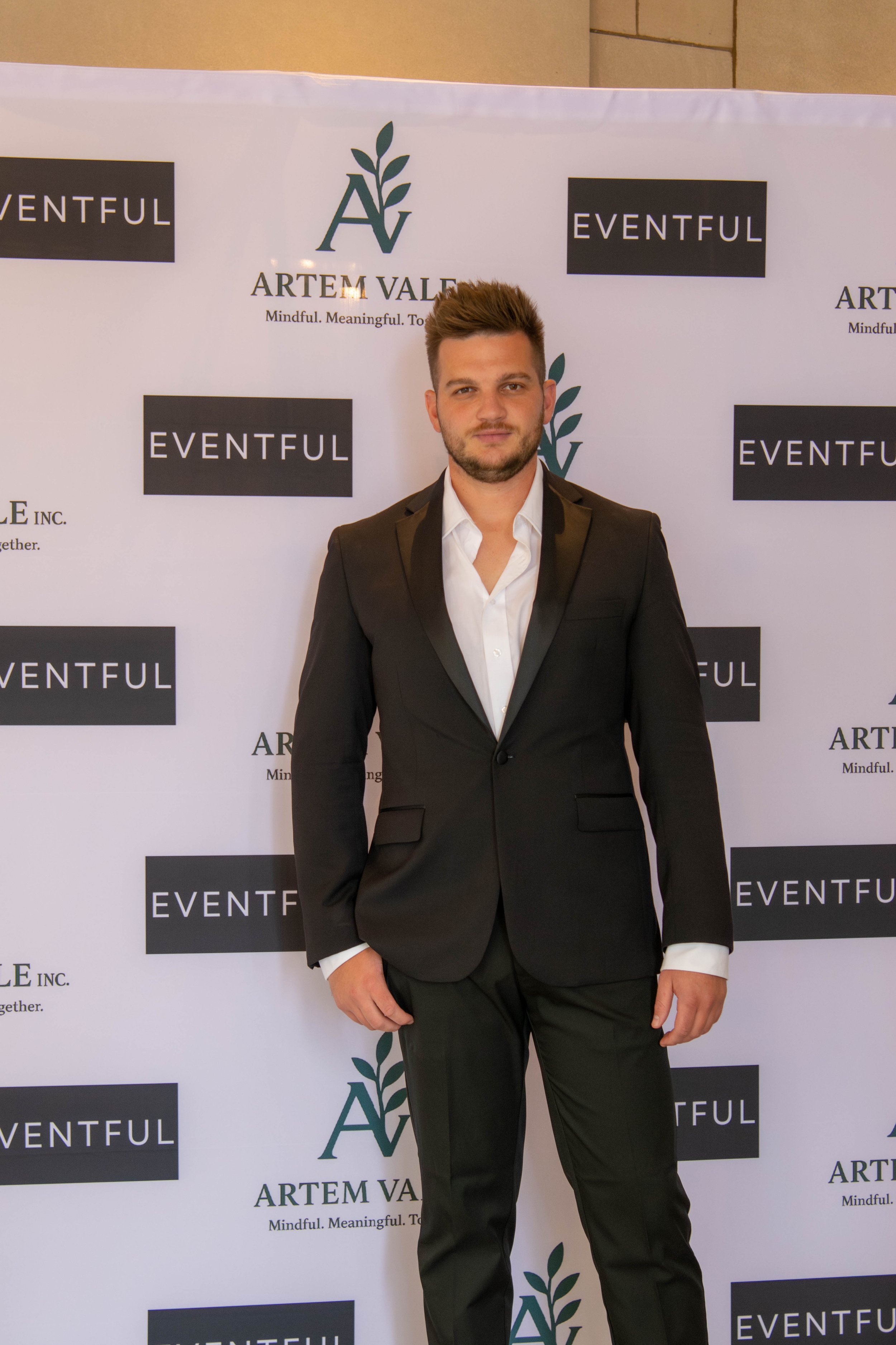 Hayden Grove in a black suit and white shirt standing in front of a backdrop at Artem Vale event in Atlanta, GA.