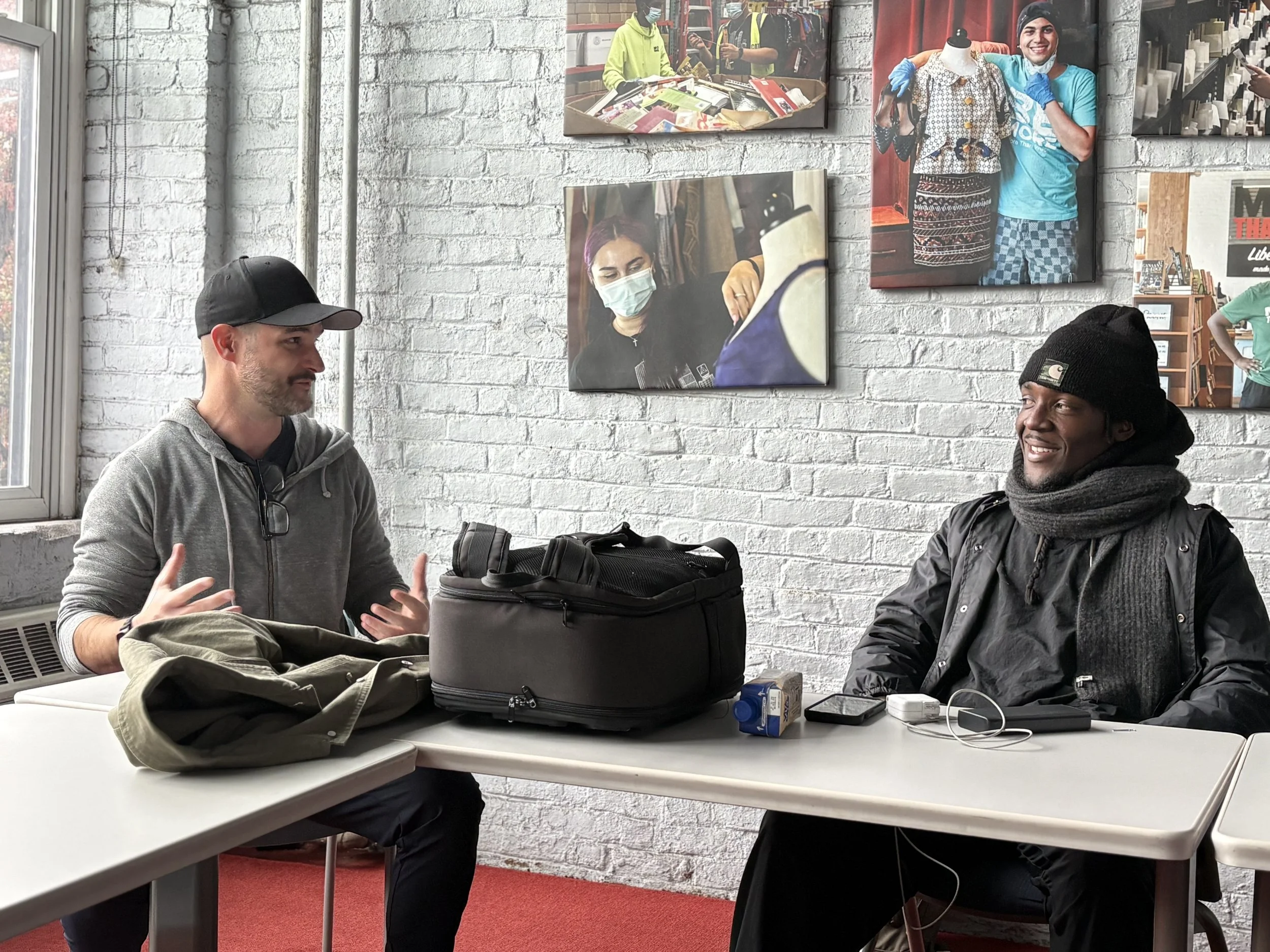 Two men are sitting at a table engaged in a conversation. The man on the left is wearing a black cap and gray hoodie, gesturing with his hands. The man on the right, dressed in dark clothing with a beanie and scarf, is smiling. On the table, there are bags, a small carton, a phone, and some electronic accessories. The background features a white brick wall with framed photographs of people working and posing.