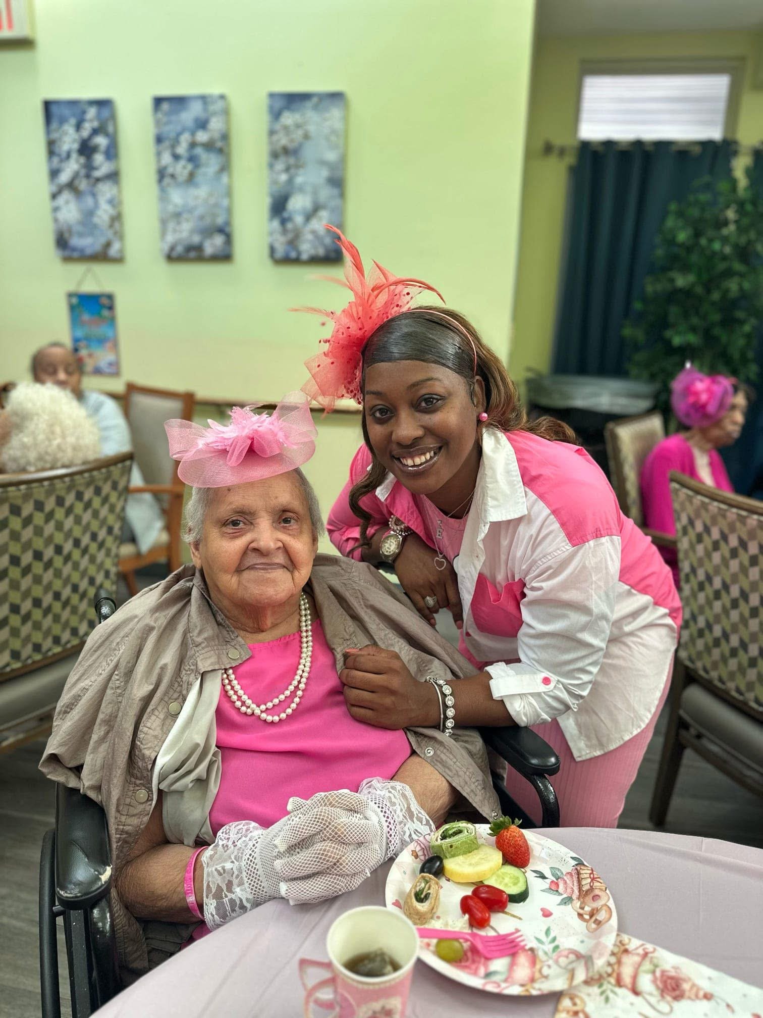 A young woman and an elderly woman celebrating at a party, both dressed in pink with hats and accessories, sitting at a table with food and drinks in a decorated room.