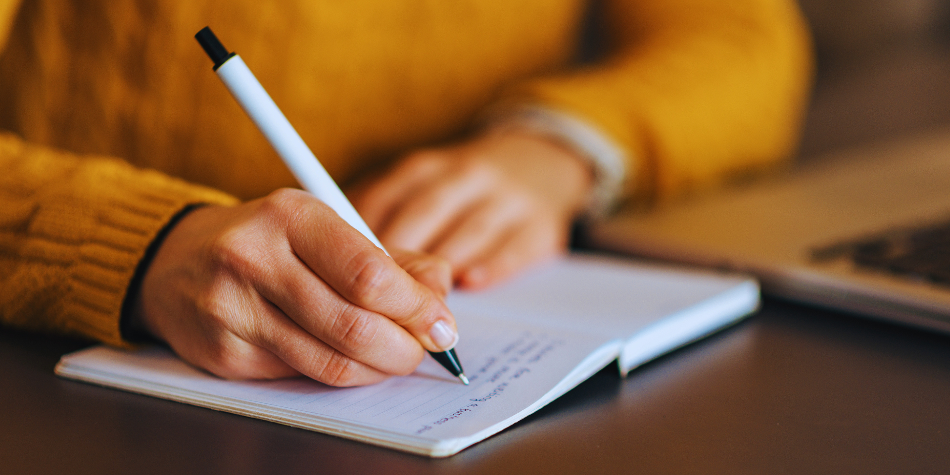 Someone in a yellow sweater writing in a notebook with a black and white pen on a brown tabletop.