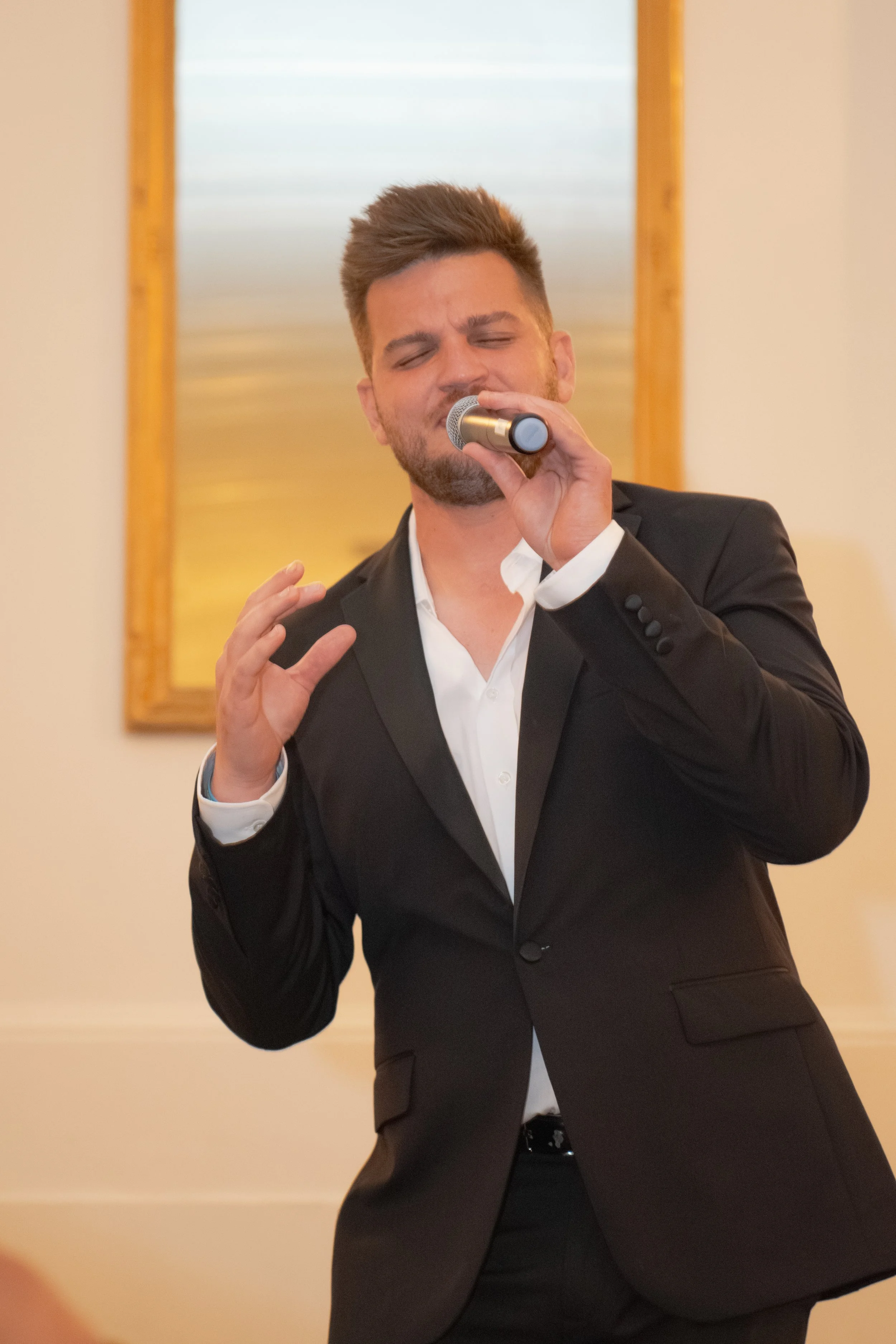 NBC the voice Hayden Grove in a black suit and white shirt singing into a microphone.