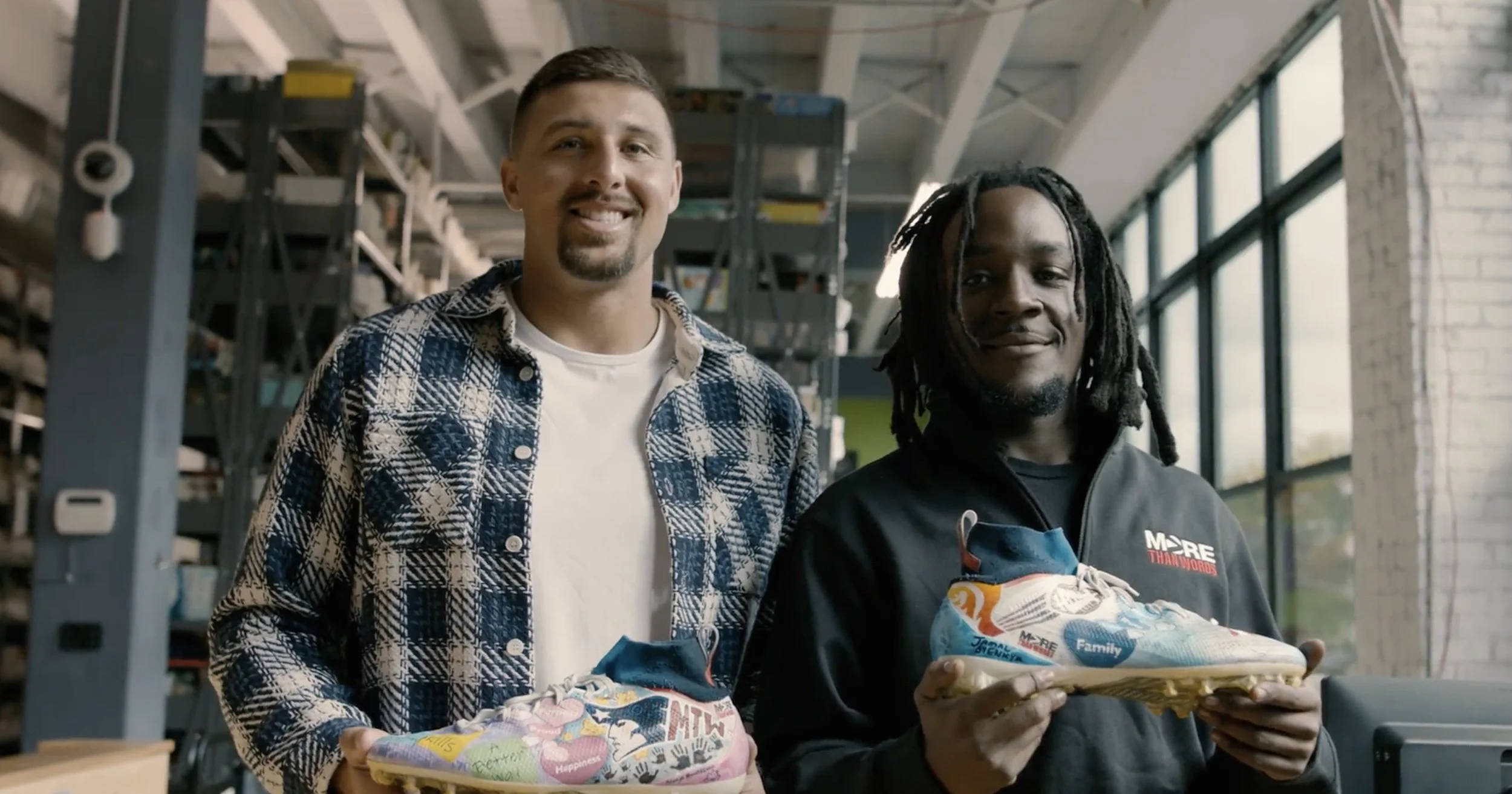 Two men standing inside a modern office space, each holding a colorful sneaker, smiling at the camera.